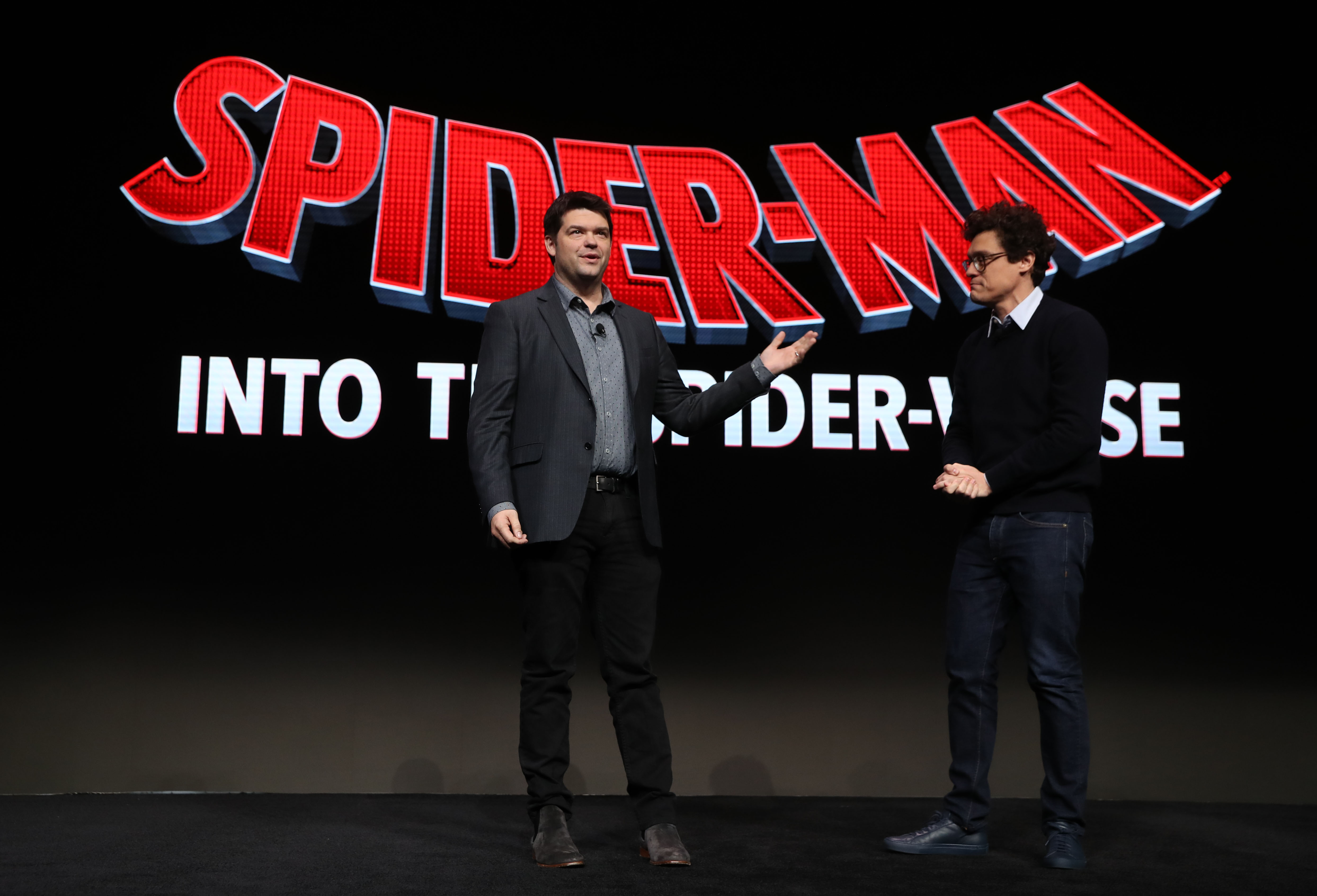 'Spider-Man: Across the Spider-Verse,' which is delayed, writers Christopher Miller and Phil Lord speak onstage. Miller wears a dark gray suit over a light gray button-up shirt and black pants. Lord wears a black sweater over a white collared shirt and jeans.