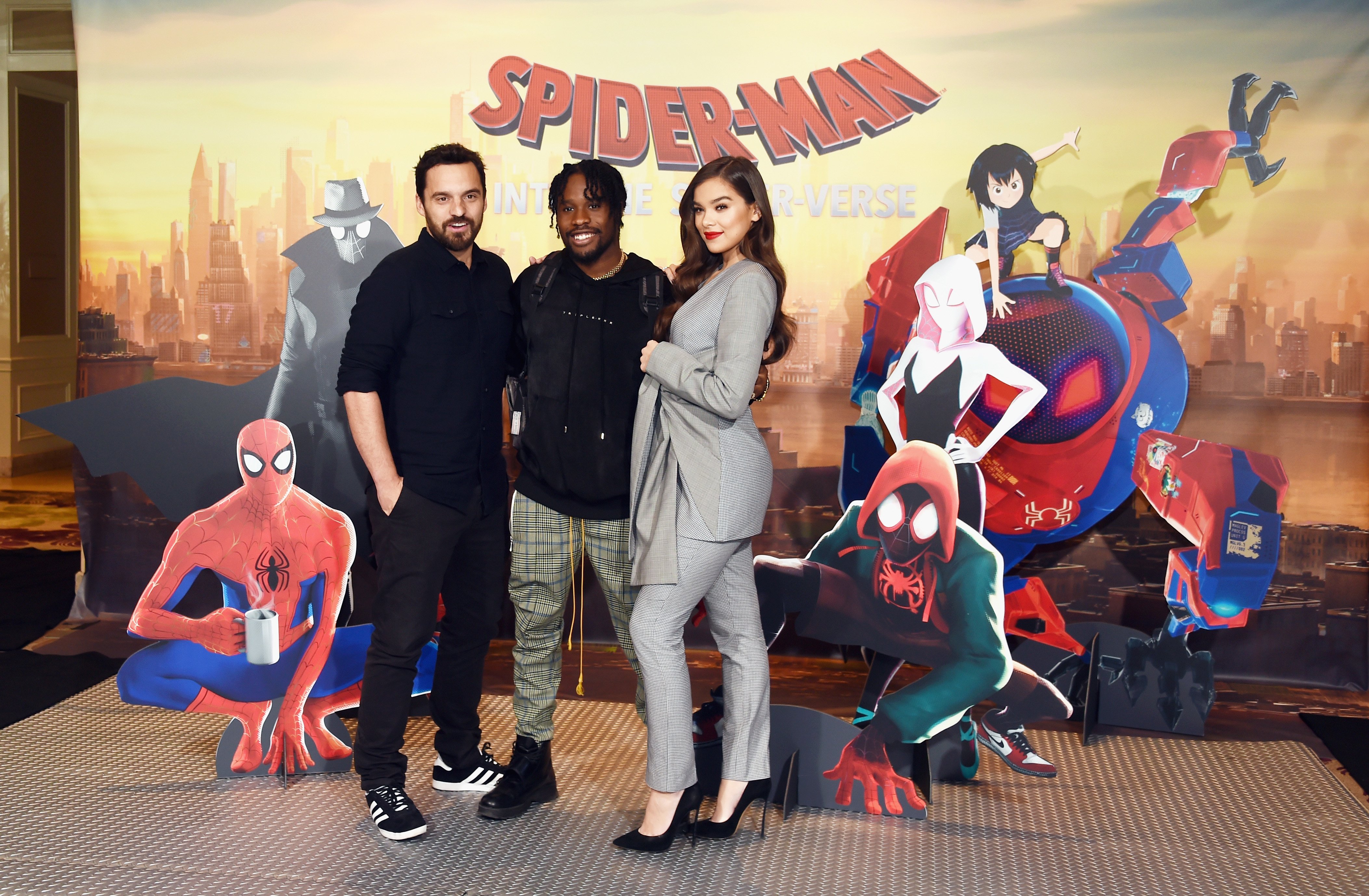 'Spider-Man: Into the Spider-Verse' stars Jake Johnson, Shameik Moore, and Hailee Steinfeld pose for pictures in front of a 3-D poster for the movie. Johnson wears a black button-up shirt and black pants. Moore wears a black hoodie and light green plaid pants. Steinfeld wears a gray suit.
