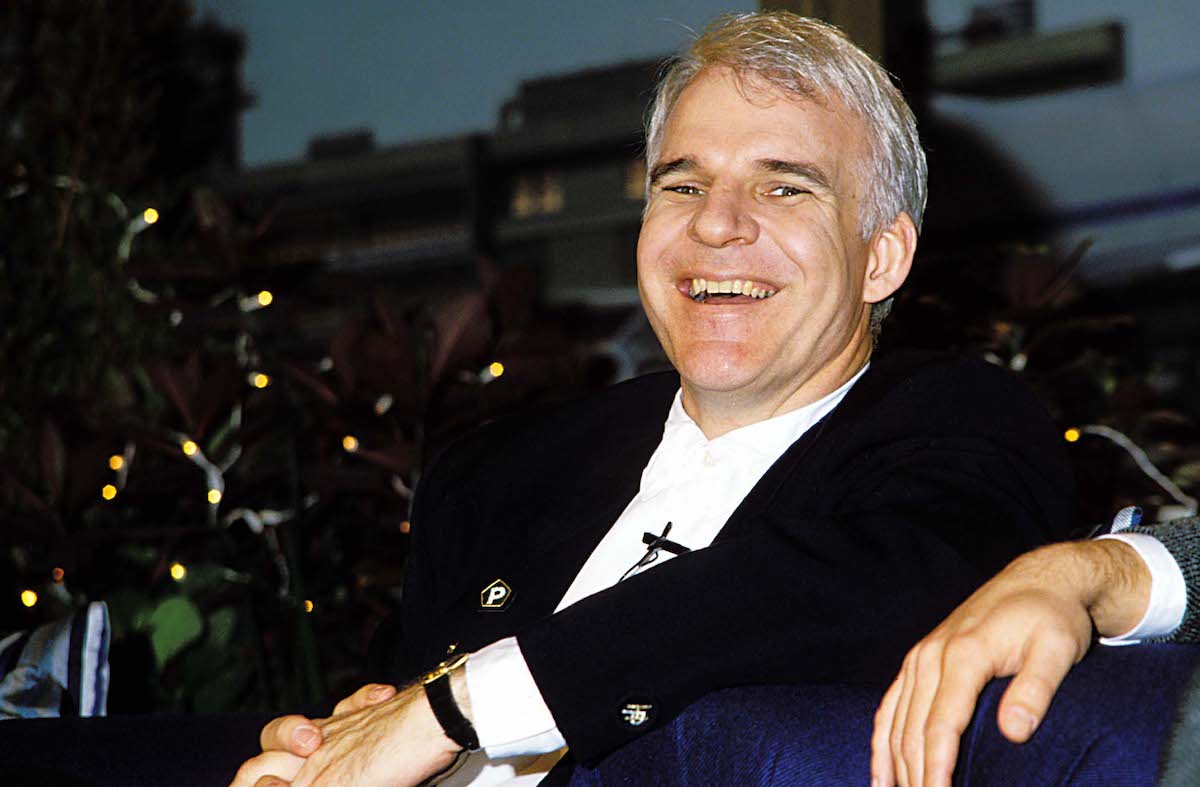 Steve Martin wears a dark jacket and laughs