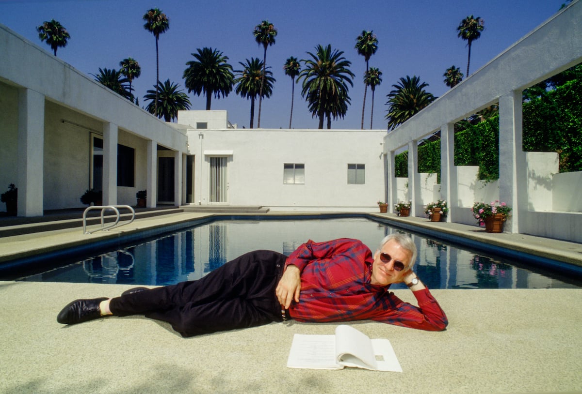 Steve Martin wears a red shirt and dark pants as he lays near a pool