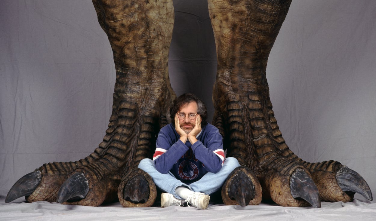 Steven Spielberg sits between a pair of prop dinosaur legs in a photo shoot promoting 'Jurassic Park'