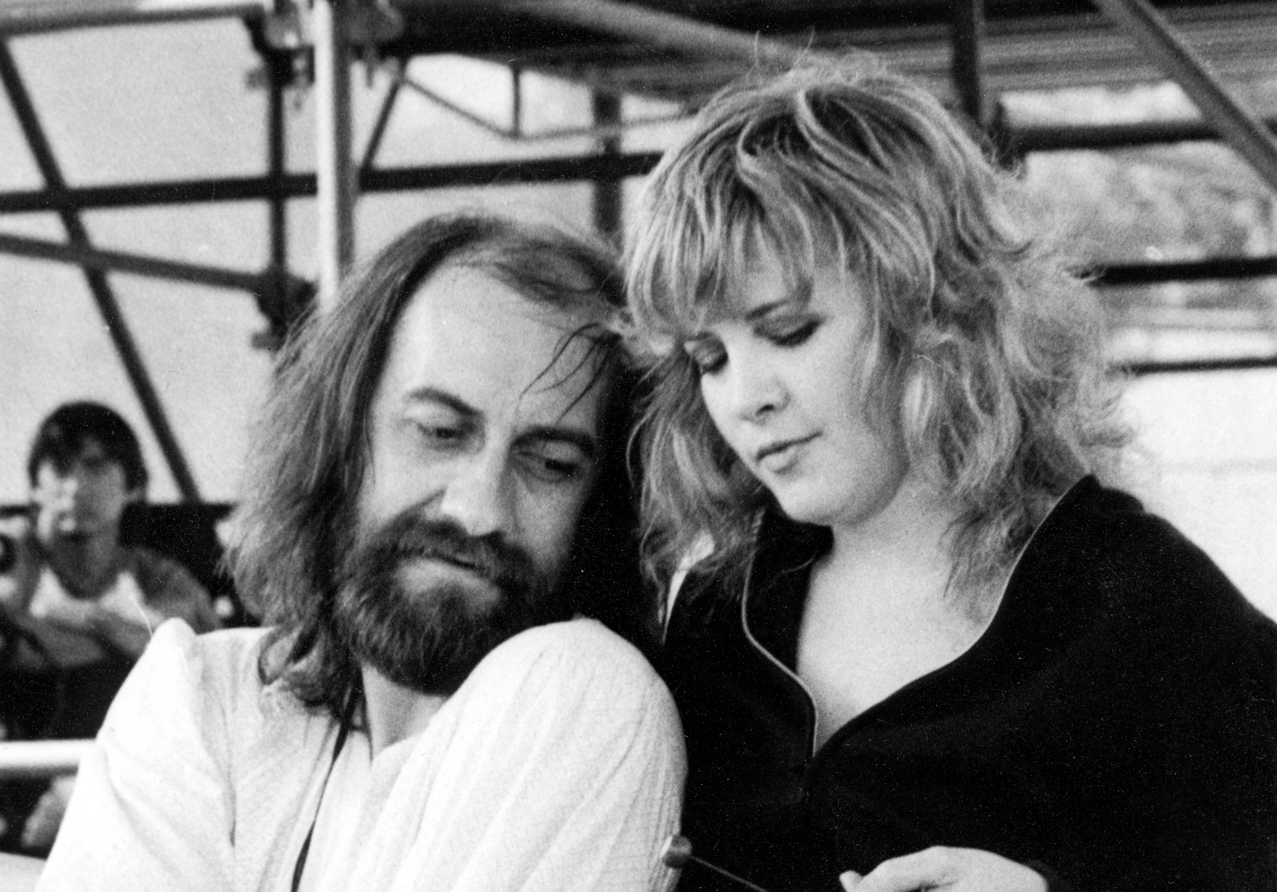 Mick Fleetwood sits and wears a white shirt. Stevie Nicks wears a black shirt and stands behind him.