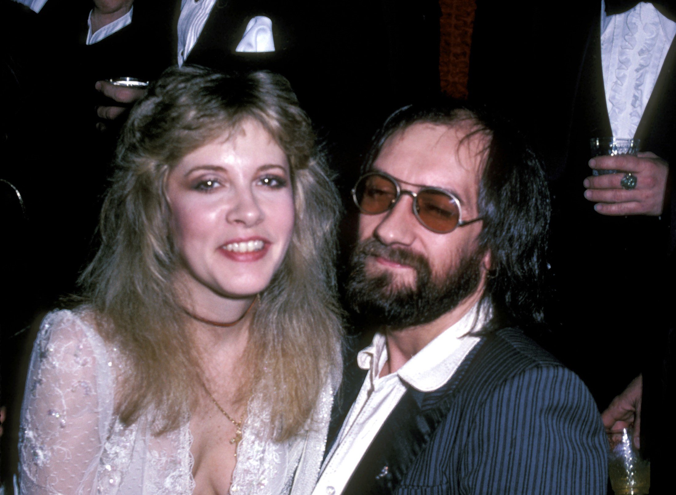 Stevie Nicks wears a white shirt and sits on Mick Fleetwood's lap. He wears sunglasses and a suit.