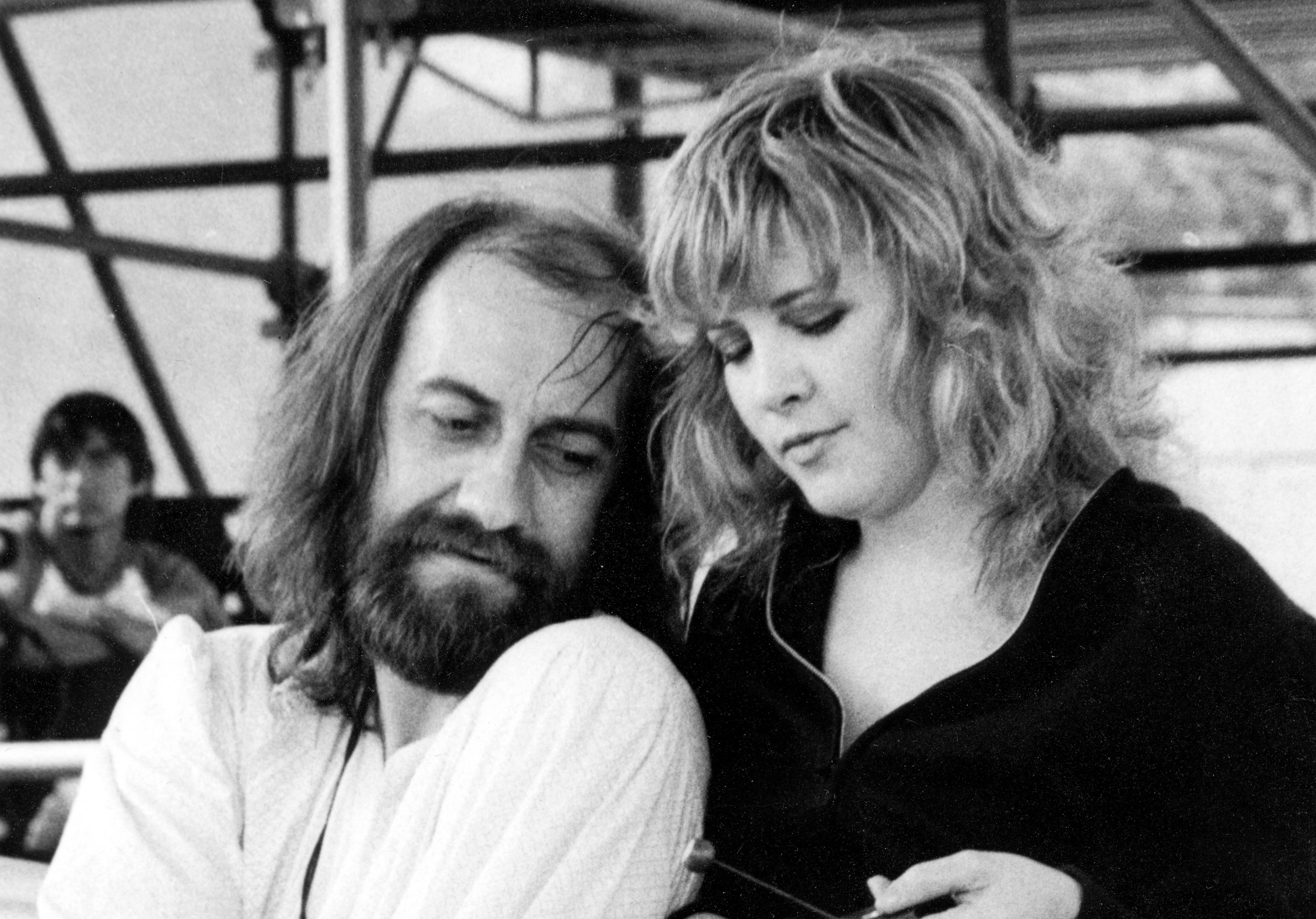 Mick Fleetwood sits at his drum set and Stevie Nicks stands behind him.