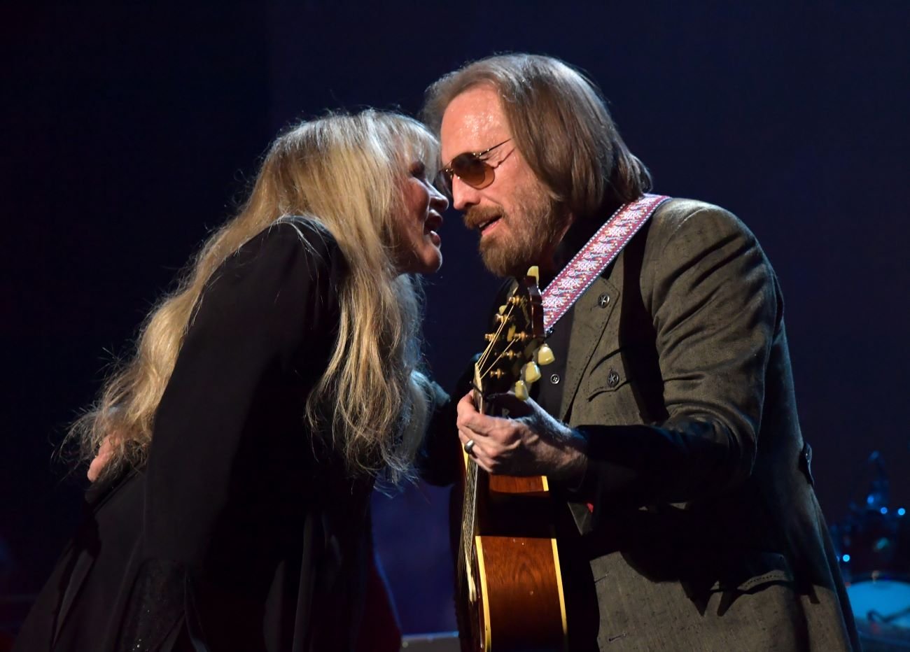 Stevie Nicks talks into Tom Petty's ear. He holds a guitar and wears sunglasses.