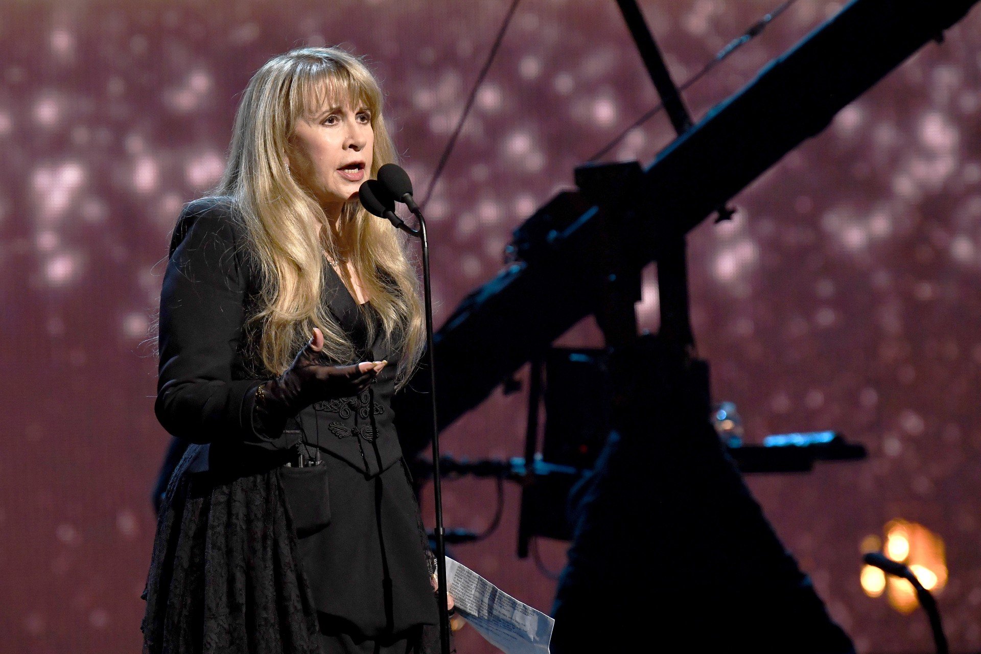 Stevie Nicks wears a long, black outfit while speaking into a microphone during a concert.