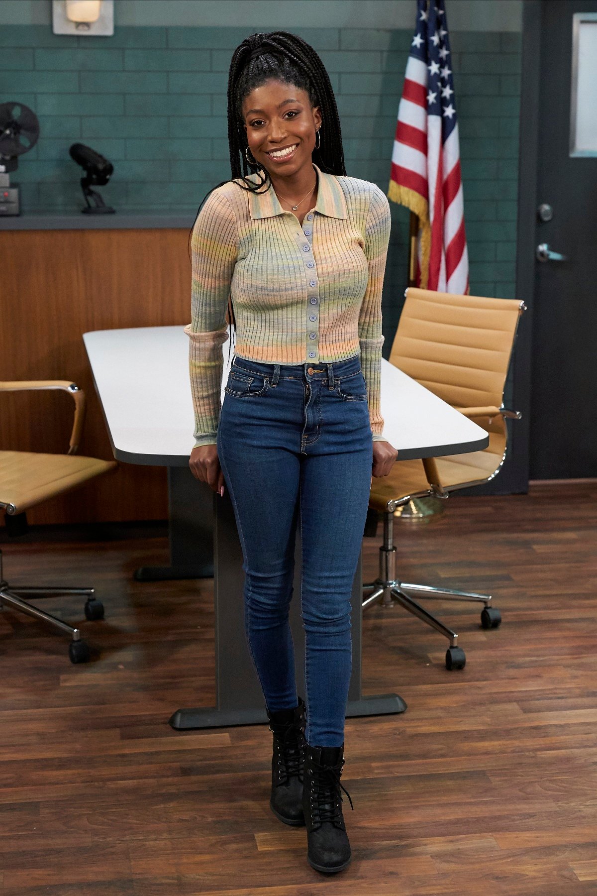 'General Hospital' actor Tabyana Ali wearing a rainbow colored sweater and jeans; stands in the interrogation room set.