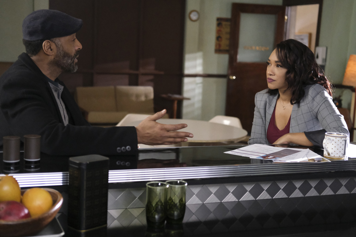 Jesse L. Martin and Candice Patton, who may appear as Joe West and Iris West-Allen in 'The Flash' Season 9, share a scene. Joe wears a black coat over a light gray shirt and a dark gray hat. Iris wears a light gray suit over a maroon shirt.