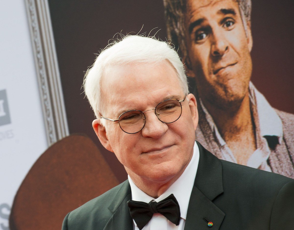 Steve Martin wears a suit and stands in front of a photo of his younger self
