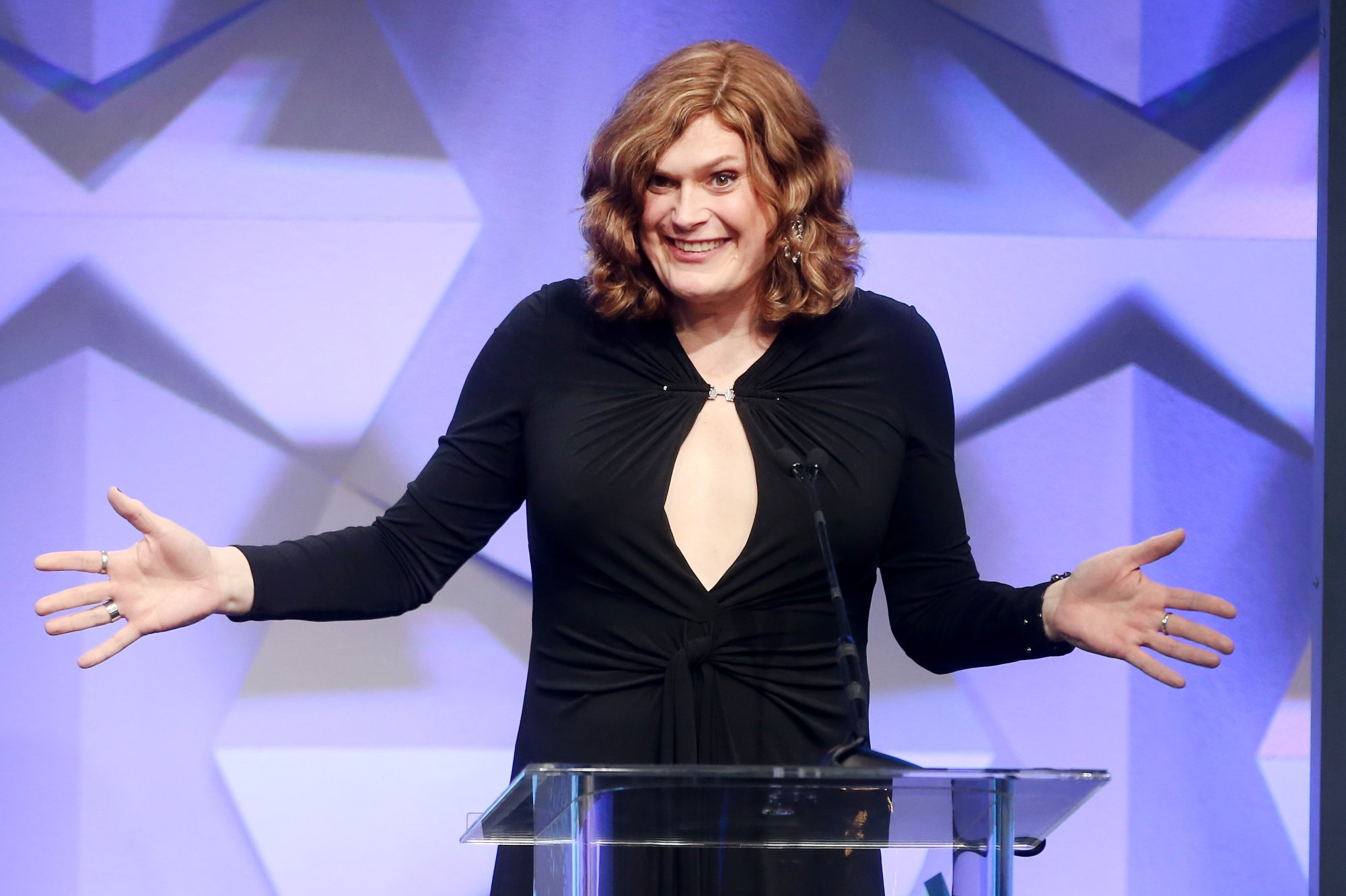 'The Matrix' filmmaker Lilly Wachowski wearing a black dress behind a glass podium with a microphone with her arms open