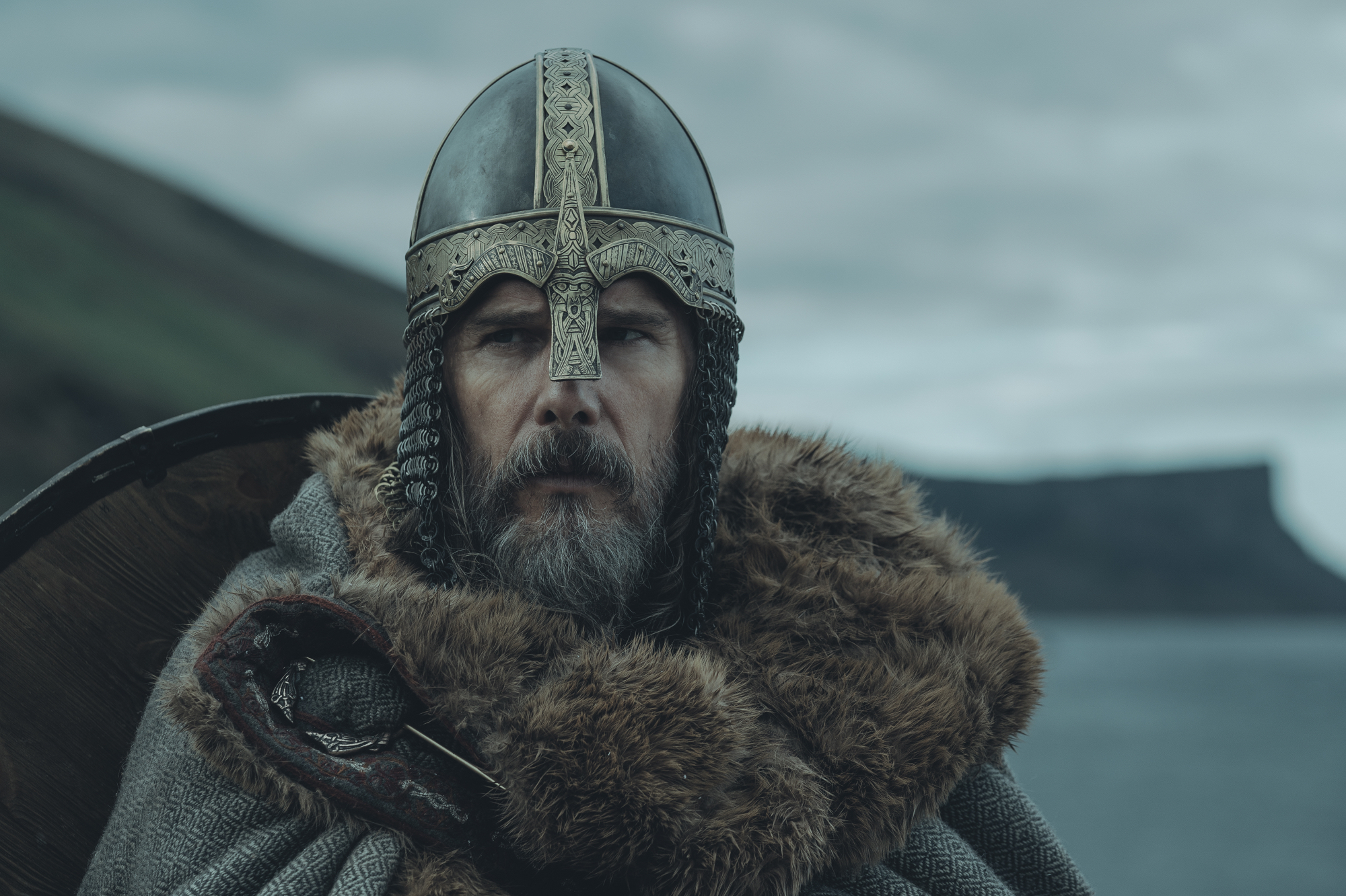 'The Northman' Ethan Hawke as King Aurvandil War-Raven wearing a helmet and coat in front of a mountain and ocean landscape
