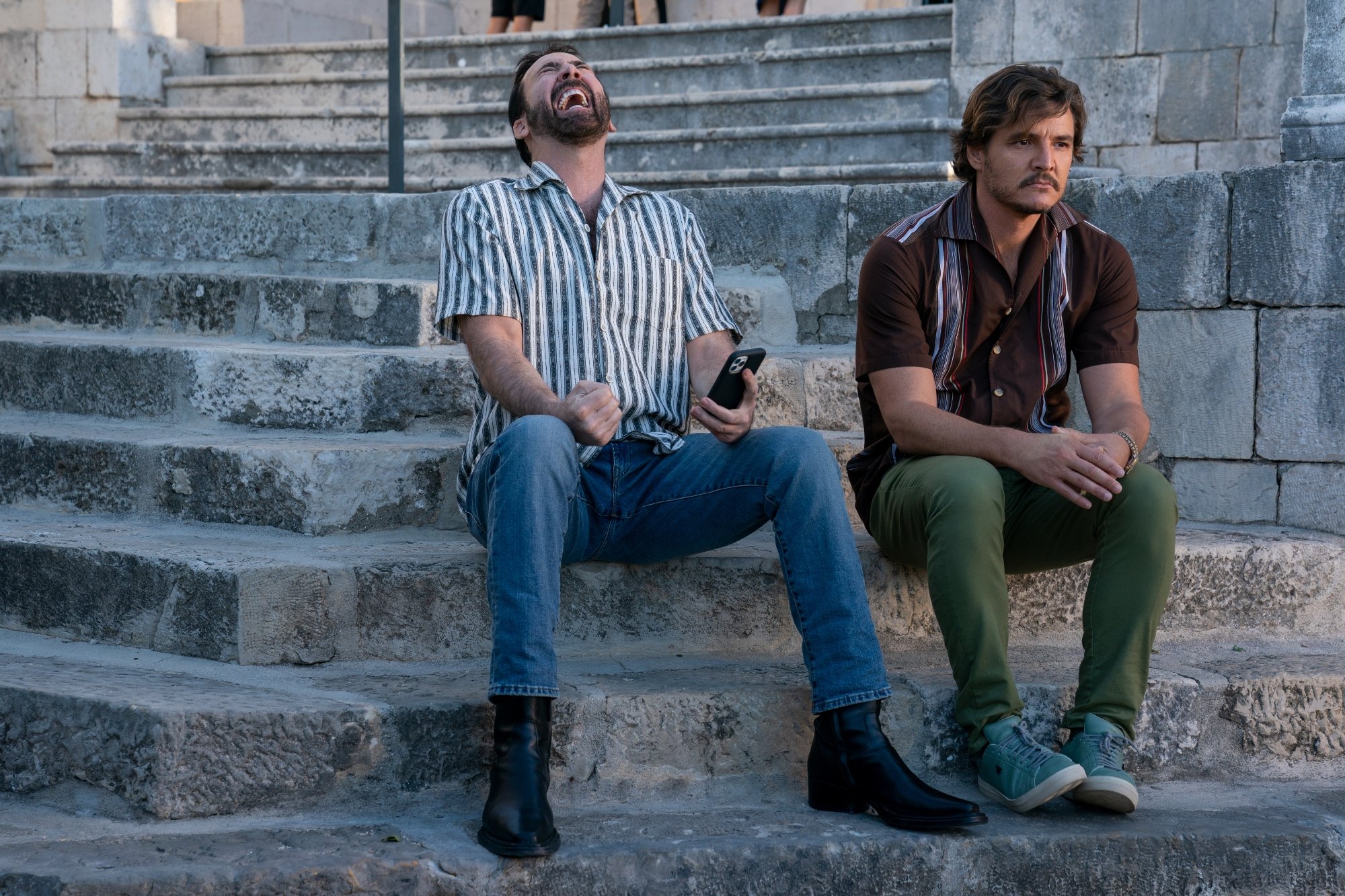 'The Unbearable Weight of Massive Talent' Nicolas Cage and Pedro Pascal as Javi Gutierrez sitting on stone stairs with Nicolas laughing and Pedro looking serious