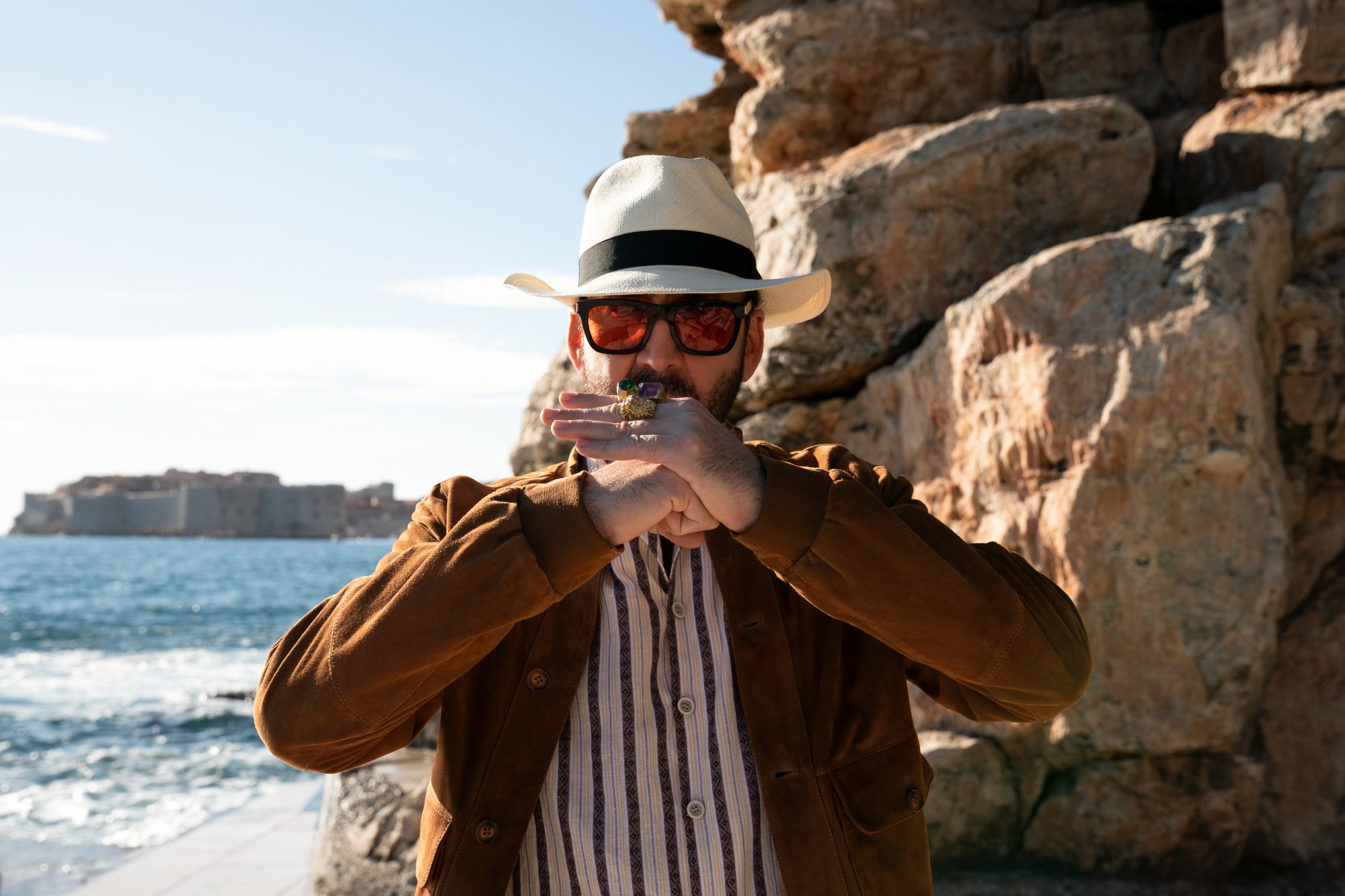 'The Unbearable Weight of Massive Talent' Nicolas Cage holding his fist in his hand, wearing a hat in front of a rock cliff and the ocean