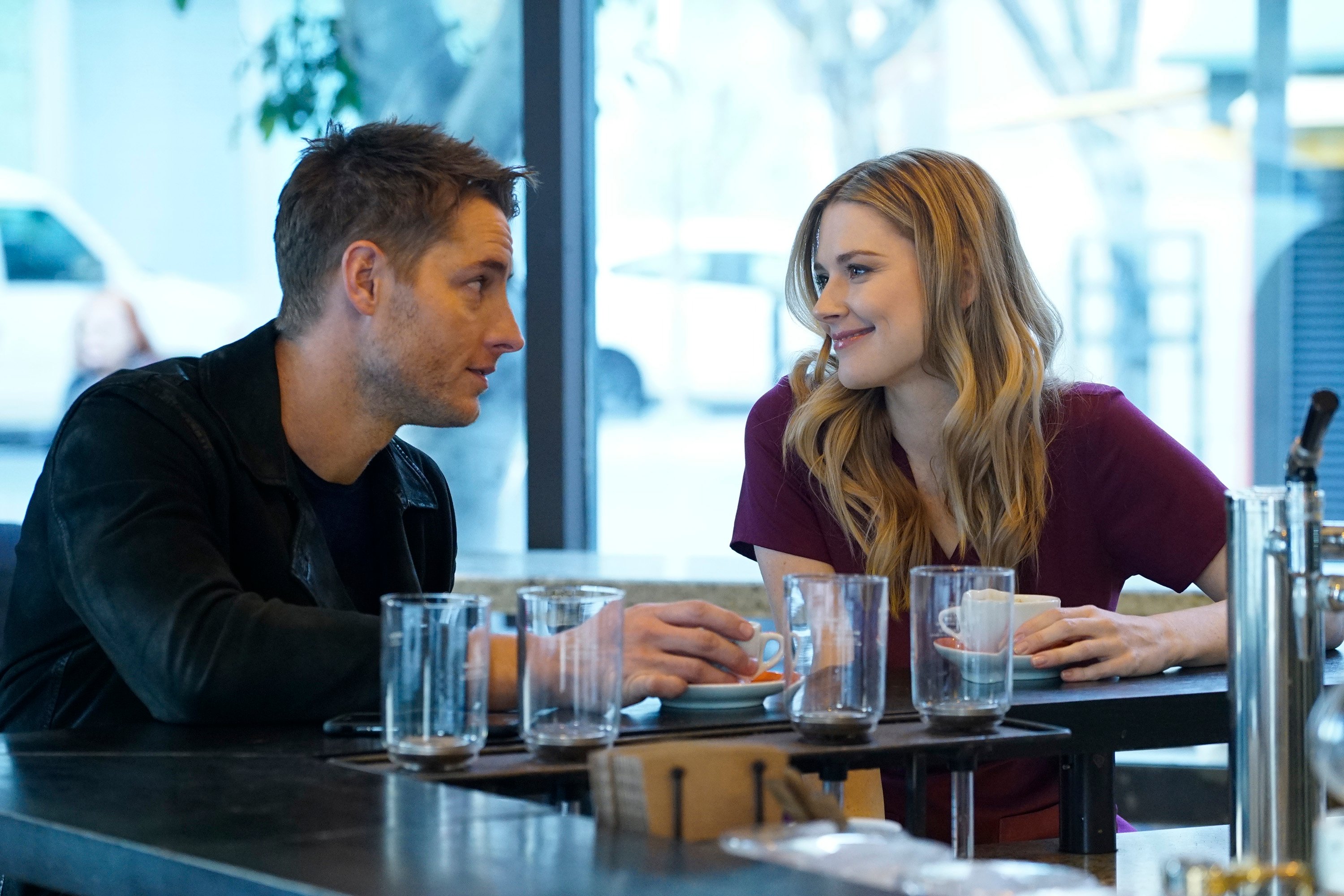 'This Is Us' stars Justin Hartley and Alexandra Breckenridge, in character as Kevin and Sophie, share a scene at a cafe. Kevin wears a black jacket over a dark shirt. Sophie wears maroon nurse scrubs.