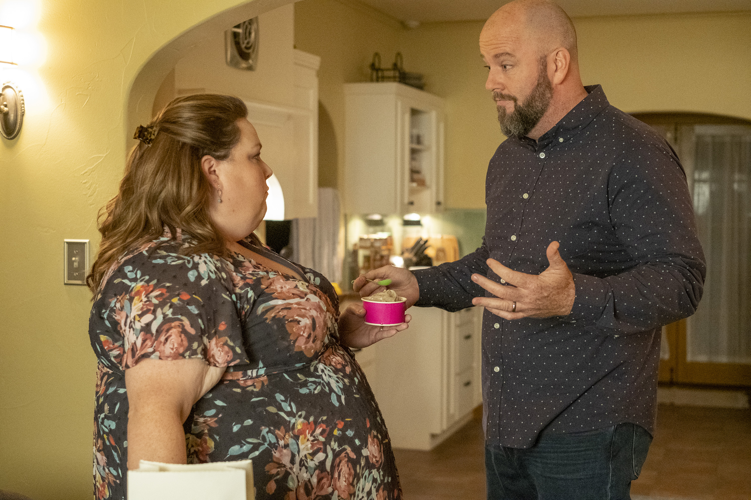 Chrissy Metz and Chris Sullivan, in character as Kate and Toby, share a scene in 'This Is Us' Season 6. Kate wears a dark gray dress with pink flowers and green leaves on it. Toby wears a black button-up shirt with white polka dots over a pair of jeans.