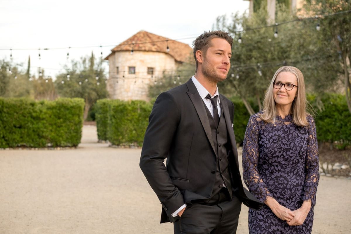 'This Is Us' Season 6 Episode 13 stars Justin Hartley and Mandy Moore, in character as Kevin and Rebecca, share a scene. Kevin wears a black suit. Rebecca wears a dark blue long-sleeved floral dress.