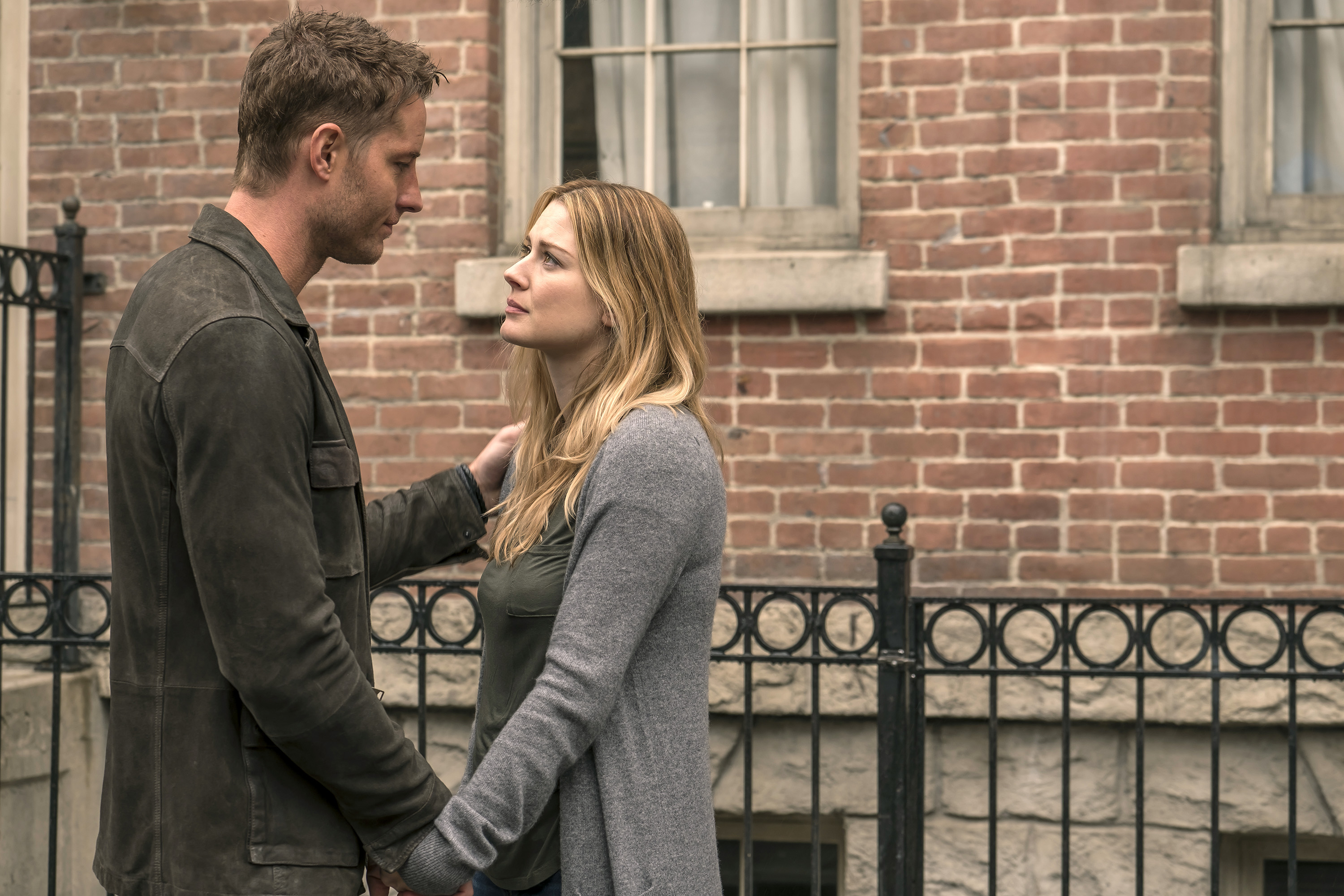 Justin Hartley and Alexandra Breckenridge, stars of 'This Is Us,' share a scene as Kevin and Sophie. Kevin, who is holding Sophie's hand and places his other hand on her shoulder, wears a dark gray jacket. Sophie wears a light gray cardigan over a dark green shirt.