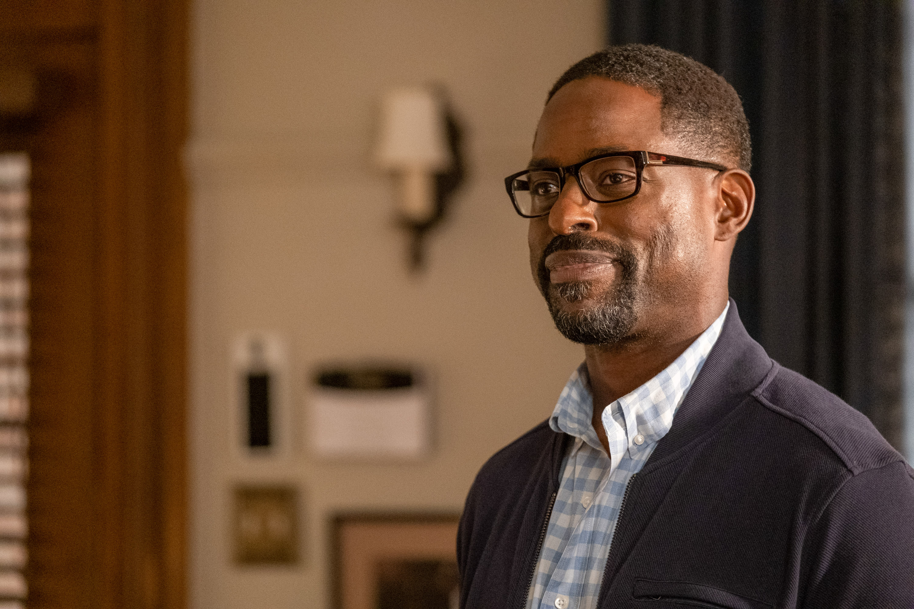 'This Is Us' star Sterling K. Brown, in character as Randall Pearson, wears a dark blue jacket over a light blue plaid button-up shirt and black-framed glasses.