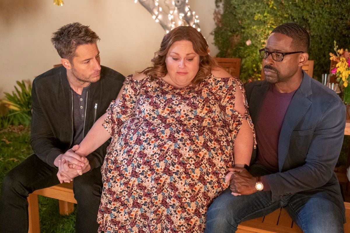 Justin Hartley, Chrissy Metz, and Sterling K. Brown, in character as Kevin, Kate, and Randall, share a scene in 'This Is Us' Season 6 Episode 11. Kate sits between her two brothers and Kevin and Randall hold her hands. Kevin wears a black jacket over a blue shirt and black pants. Kate wears a purple, blue, yellow, and white floral dress. Randall wears a dark gray blazer over a maroon shirt and jeans.