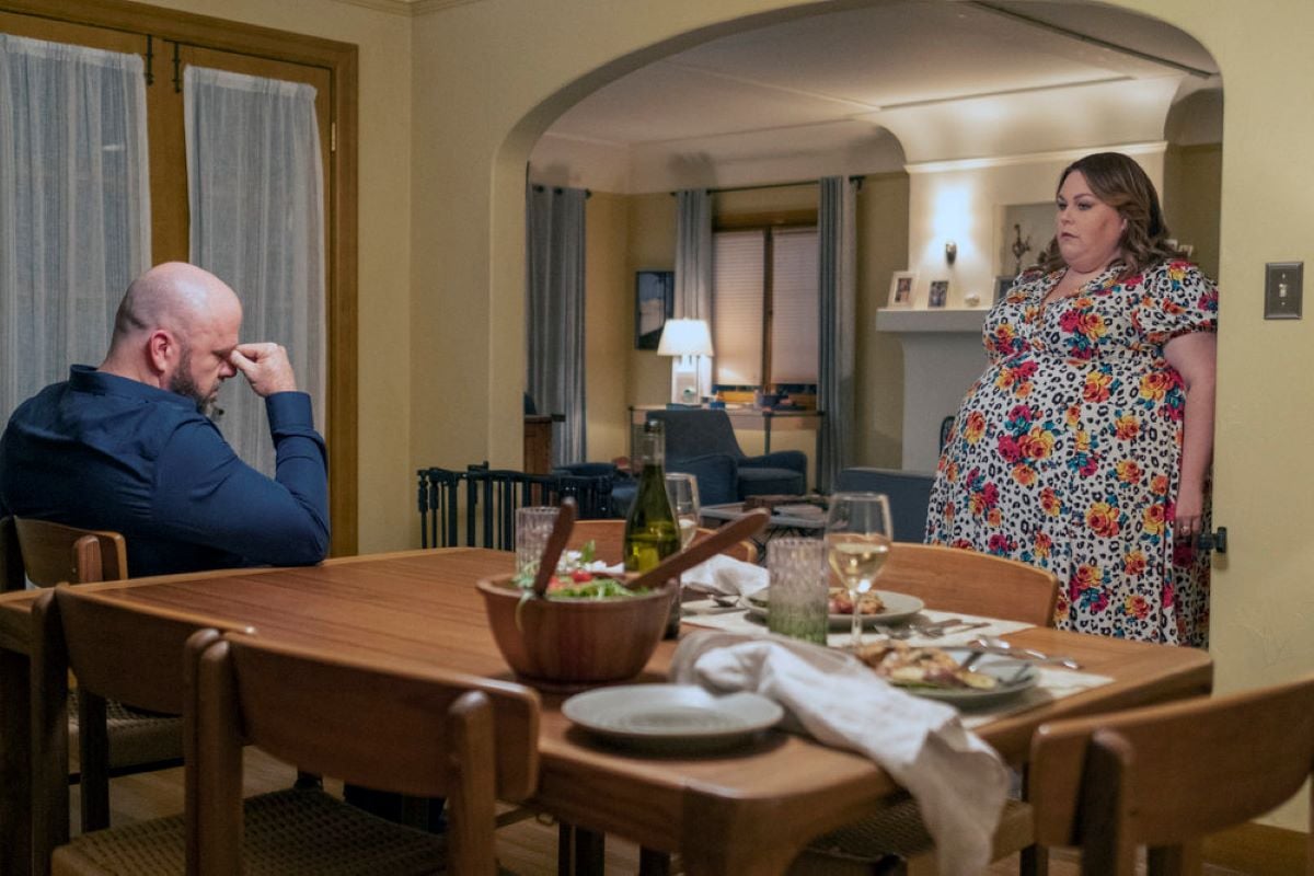 'This Is Us' Season 6 Episode 12 stars Chris Sullivan and Chrissy Metz, in character as Toby and Kate, share a scene where Toby sits at the kitchen table and Kate stands in the doorway. Toby wears a dark blue button-up shirt. Kate wears a white dress with leopard print and yellow and red roses on it.
