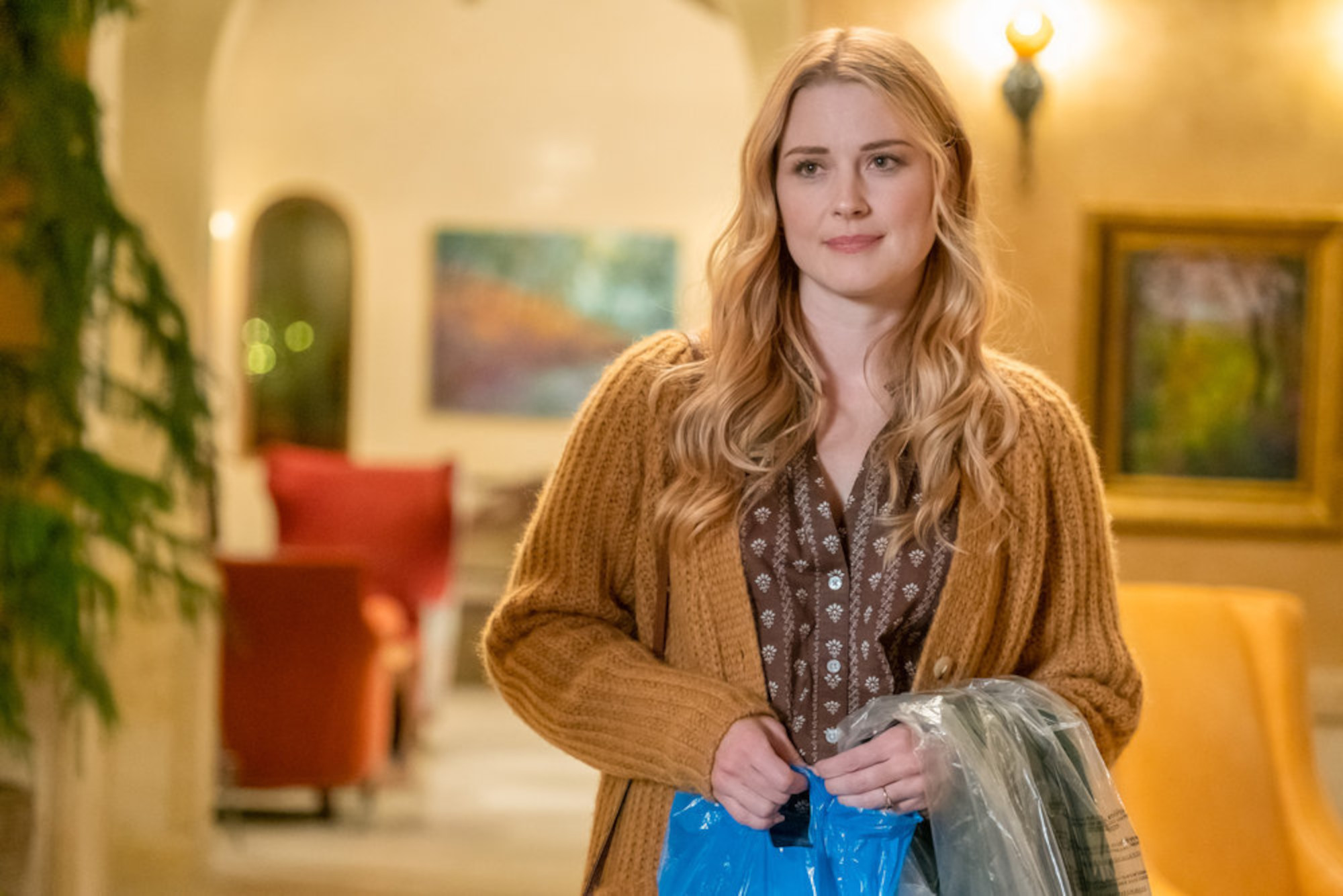 Alexandra Breckenridge as Sophie Inman in 'This Is Us' Season 6 Episode 14. She's wearing a yellow sweater, holding a blue bag, and smiling.