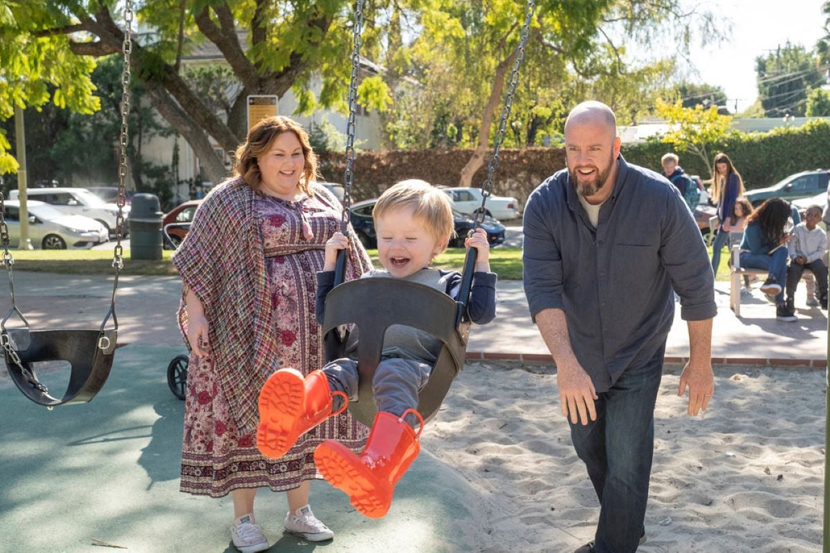 Chrissy Metz, Jonathan Kincaid, and Chris Sullivan, in character as Kate, Jack, and Toby, share a scene in 'This Is Us' Season 6 Episode 11, which airs tonight, April 5. Kate and Toby smile at Jack while pushing him on a swing. Kate wears a pink and red floral dress and a plaid shawl. Jack wears a gray and dark blue shirt, gray pants, and red rain boots. Toby wears a dark gray button-up shirt and jeans.