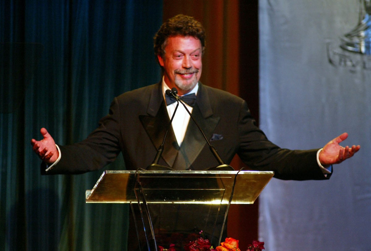 Tim Curry wears a suit and holds his arms out