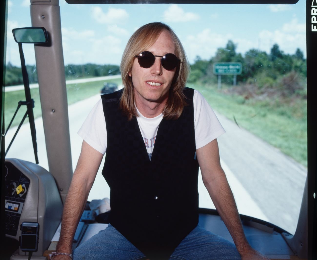 Tom Petty sits near the front window inside a bus and wears a black vest and sunglasses