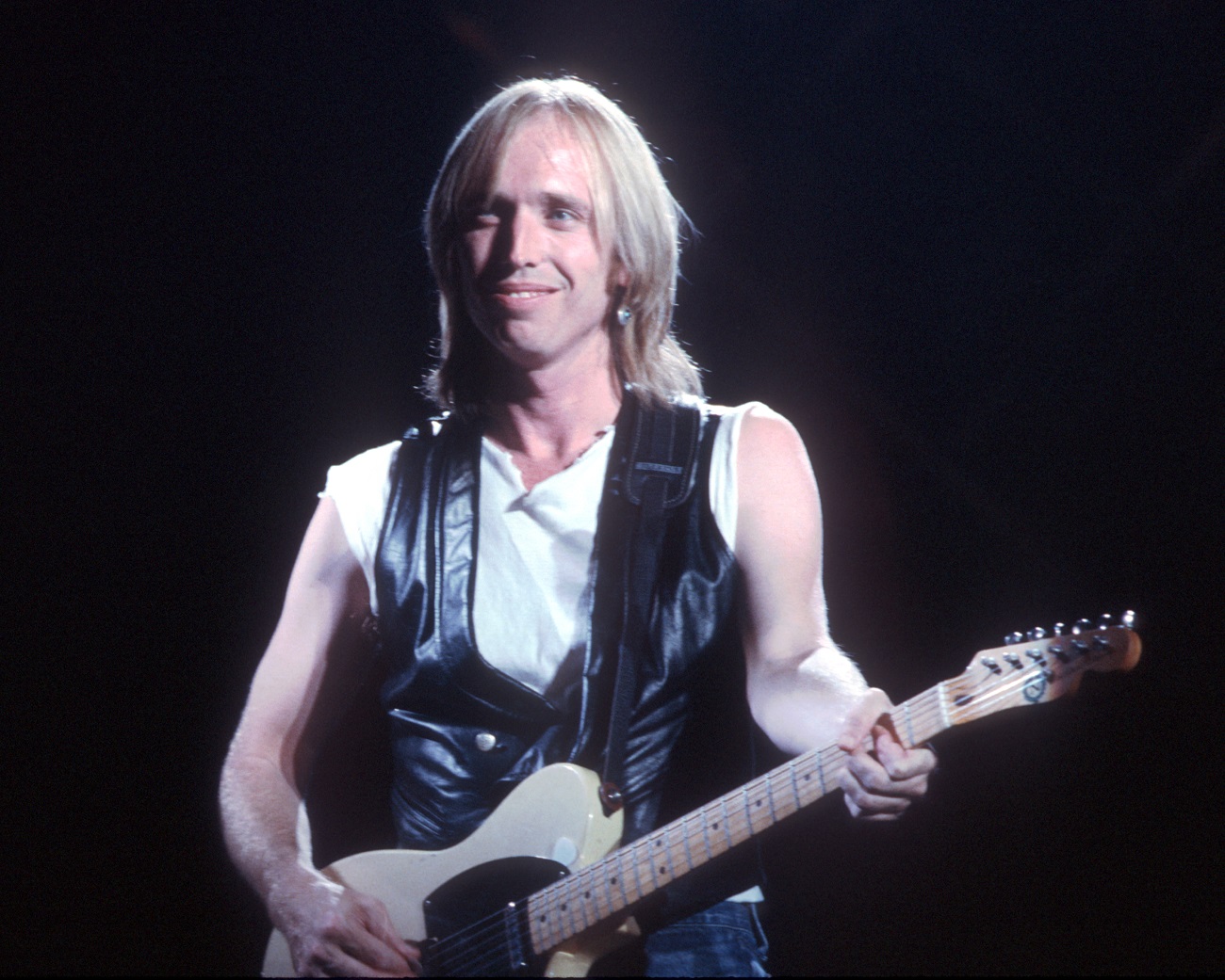 Tom Petty wears a white tank top and black vest and plays the guitar.