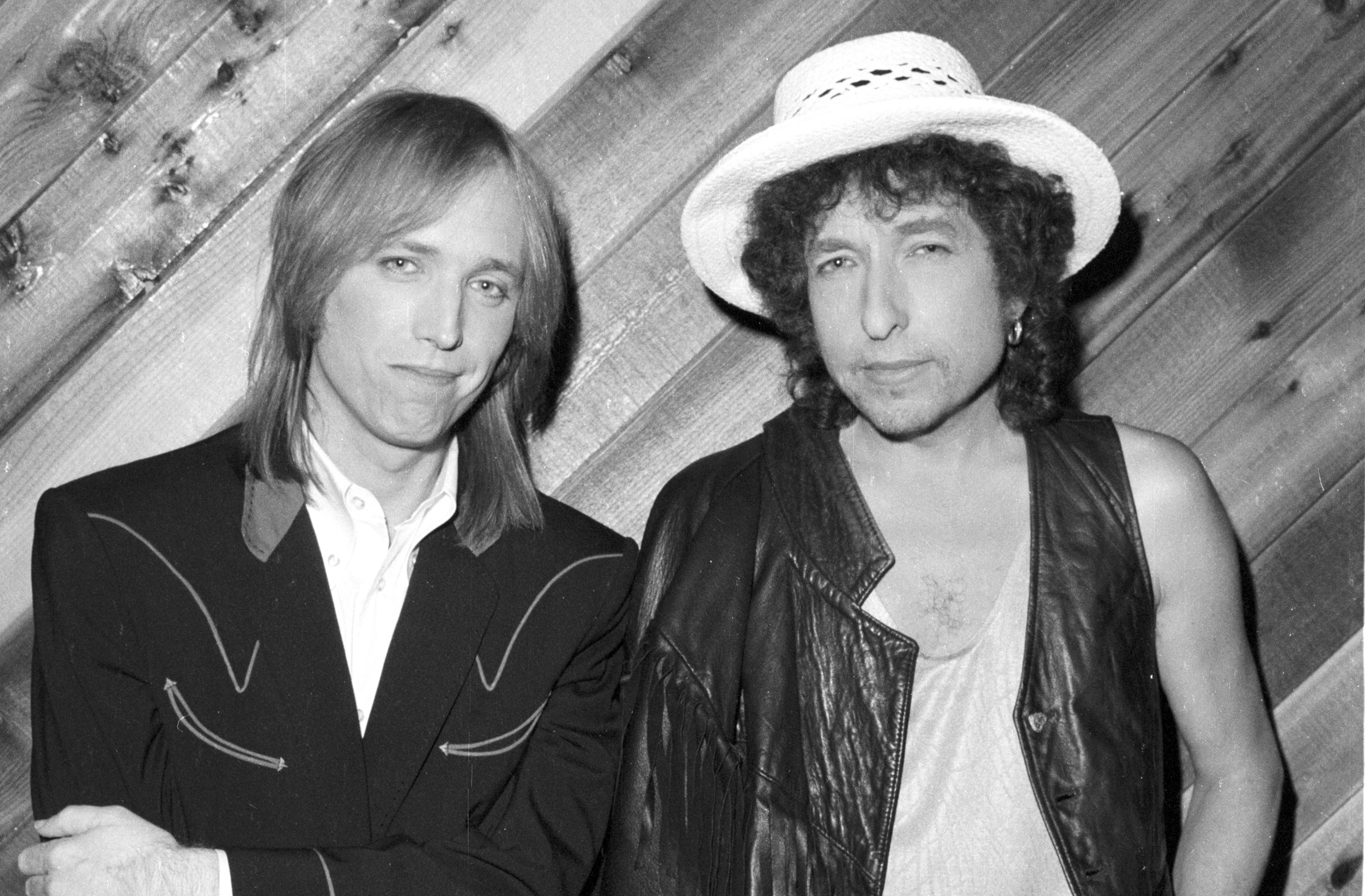 A black and white photo of Tom Petty and Bob Dylan leaning against a wood paneled wall.
