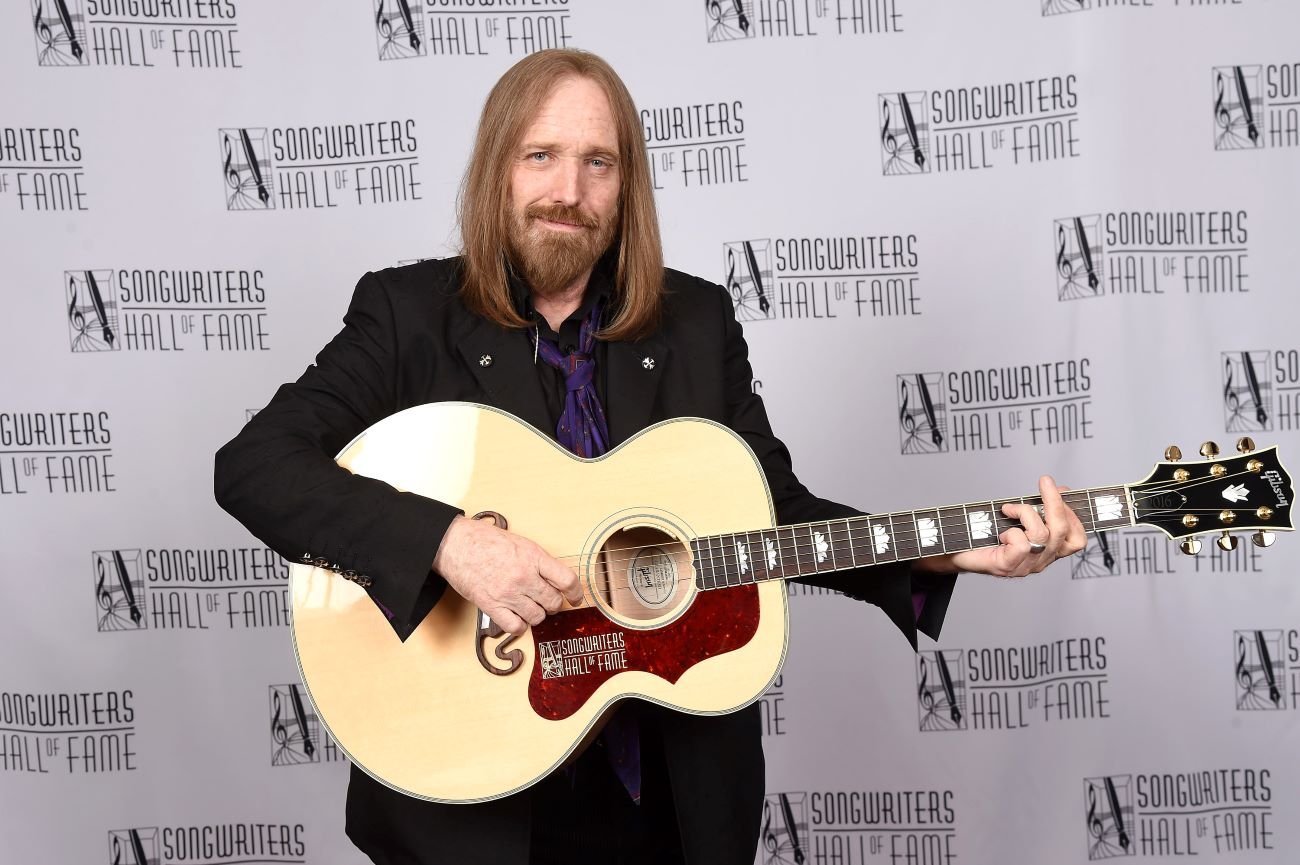 Tom Petty wears a black suit and holds a guitar. He stands in front of the Songwriter's Hall of Fame background.
