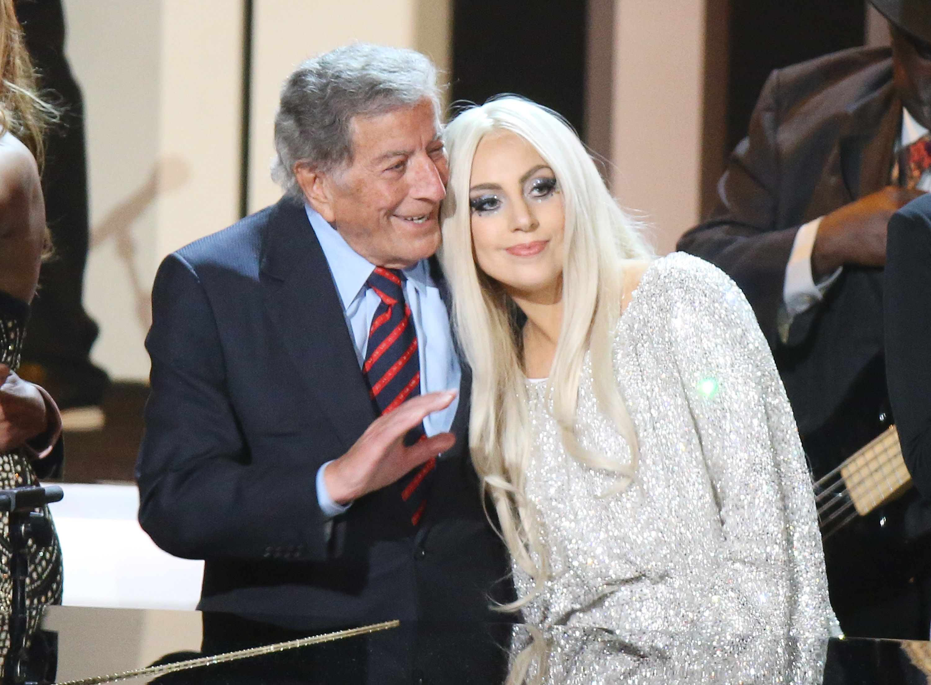 Tony Bennet and Lady Gaga stand close together on stage. He wears a suit and she wears a sparkly dress.