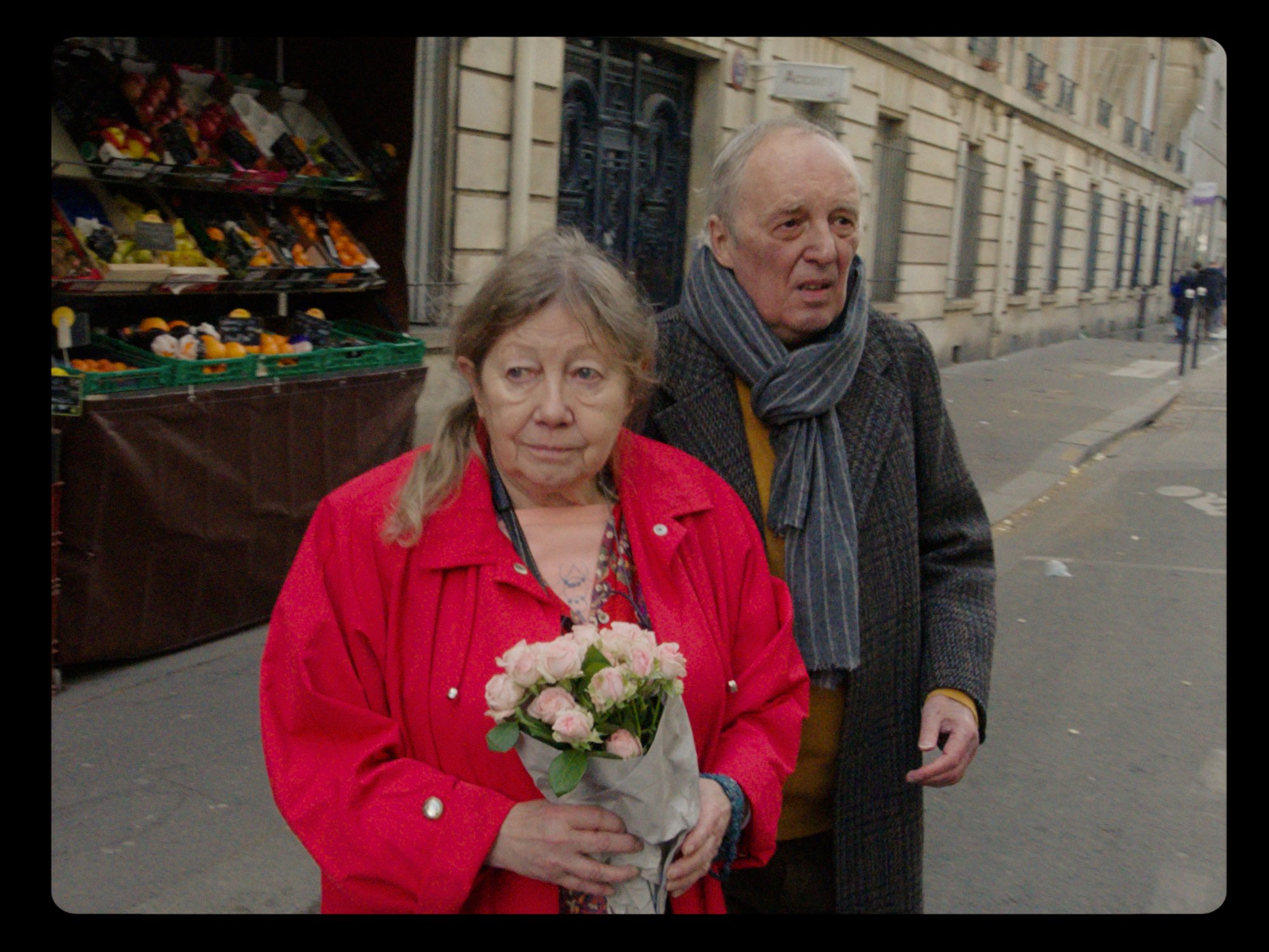 'Vortex' Françoise Lebrun as Elle and Dario Argento as Lui with Lebrun holding flowers in a red jacket walking into the street in front of a store