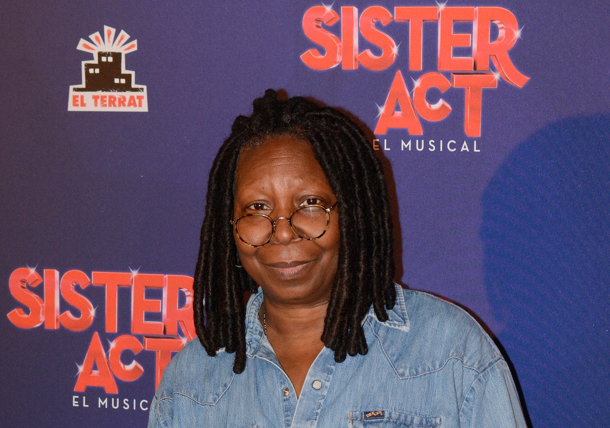 Whoopi Goldberg wears denim and poses in front of the Sister Act: The Musical logo
