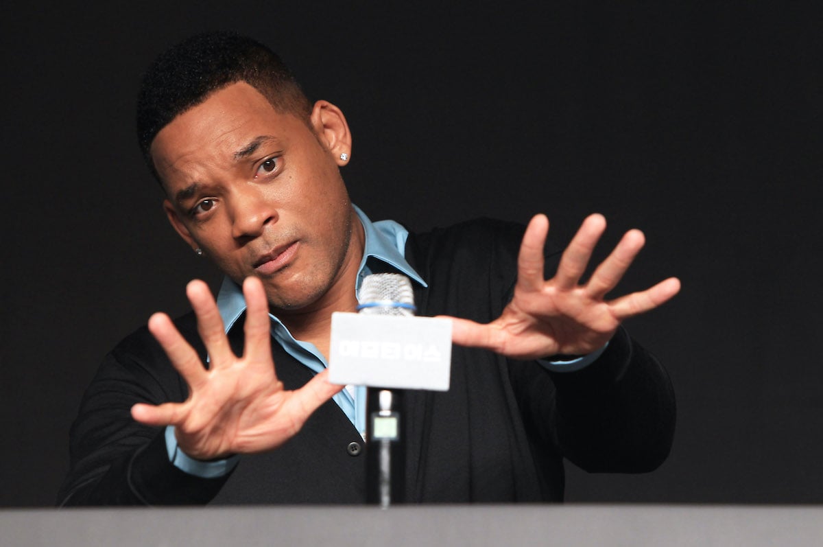 Will Smith holds his hands in front of him as he speaks at a microphone