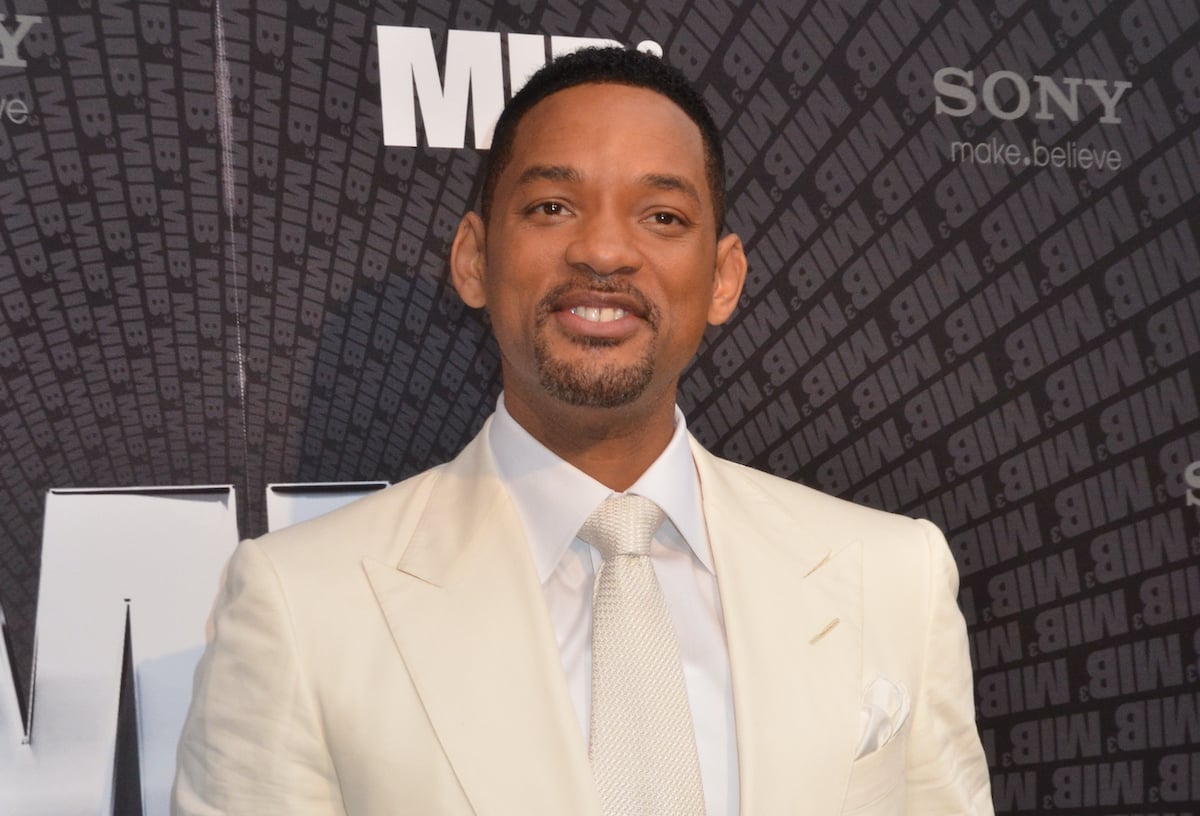 Will Smith wears a suit and poses in front of the ‘Men In Black 3’ logo