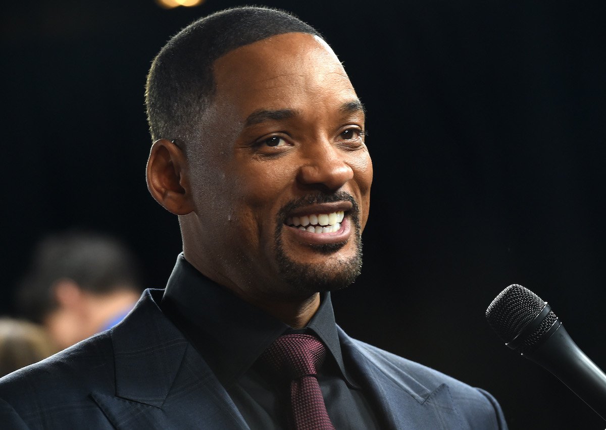 Will Smith smiles in a suit at a microphone