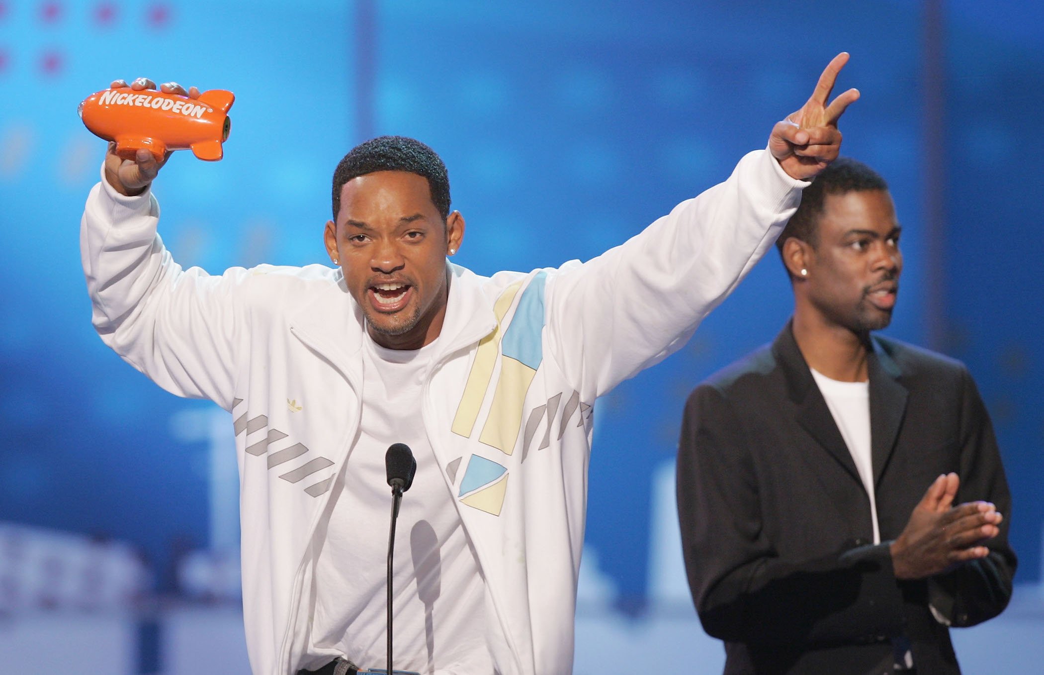 Will Smith at the Kids Choice Awards accepting an award with Chris Rock in the background. Will Smith is excitedly standing at the podium holding his award.