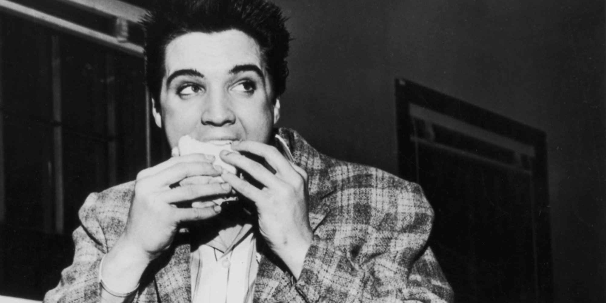 Elvis Presley enjoys a sandwich in a black and white photo.