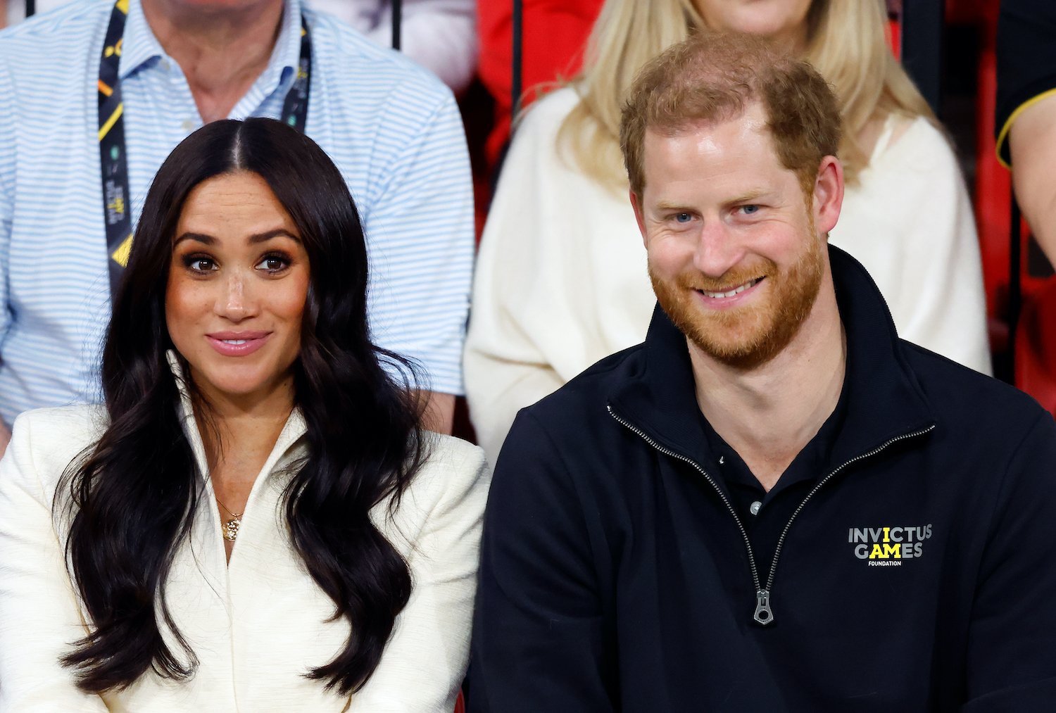 Meghan Markle wears a white top and smiles alongside Prince Harry smiling and wearing a dark half zip pullover at the Invictus Games