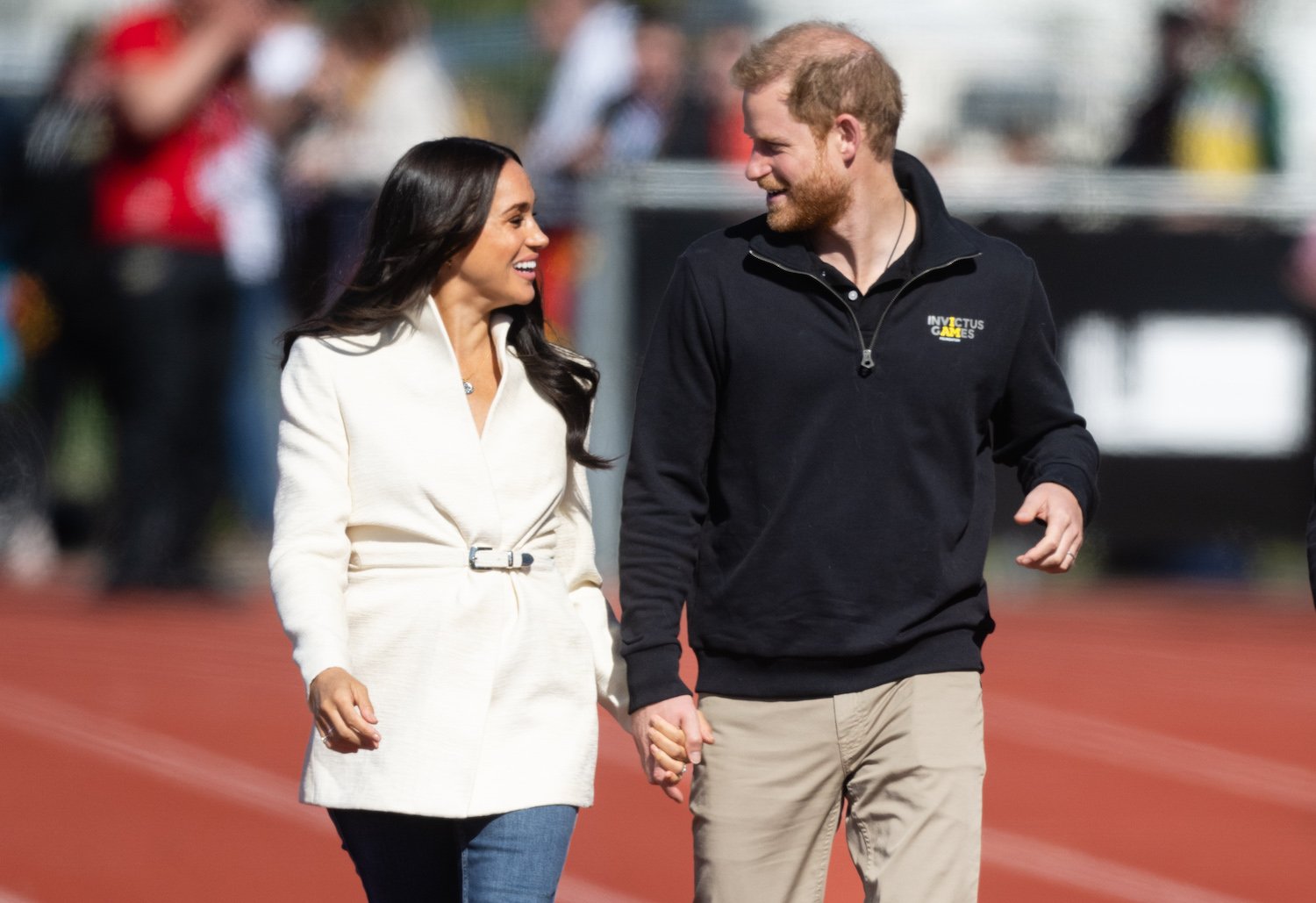 Meghan Markle wears a white coat and jeans and smiles at Prince Harry, wearing khaki pants and a dark half-zip top at the Invictus Games