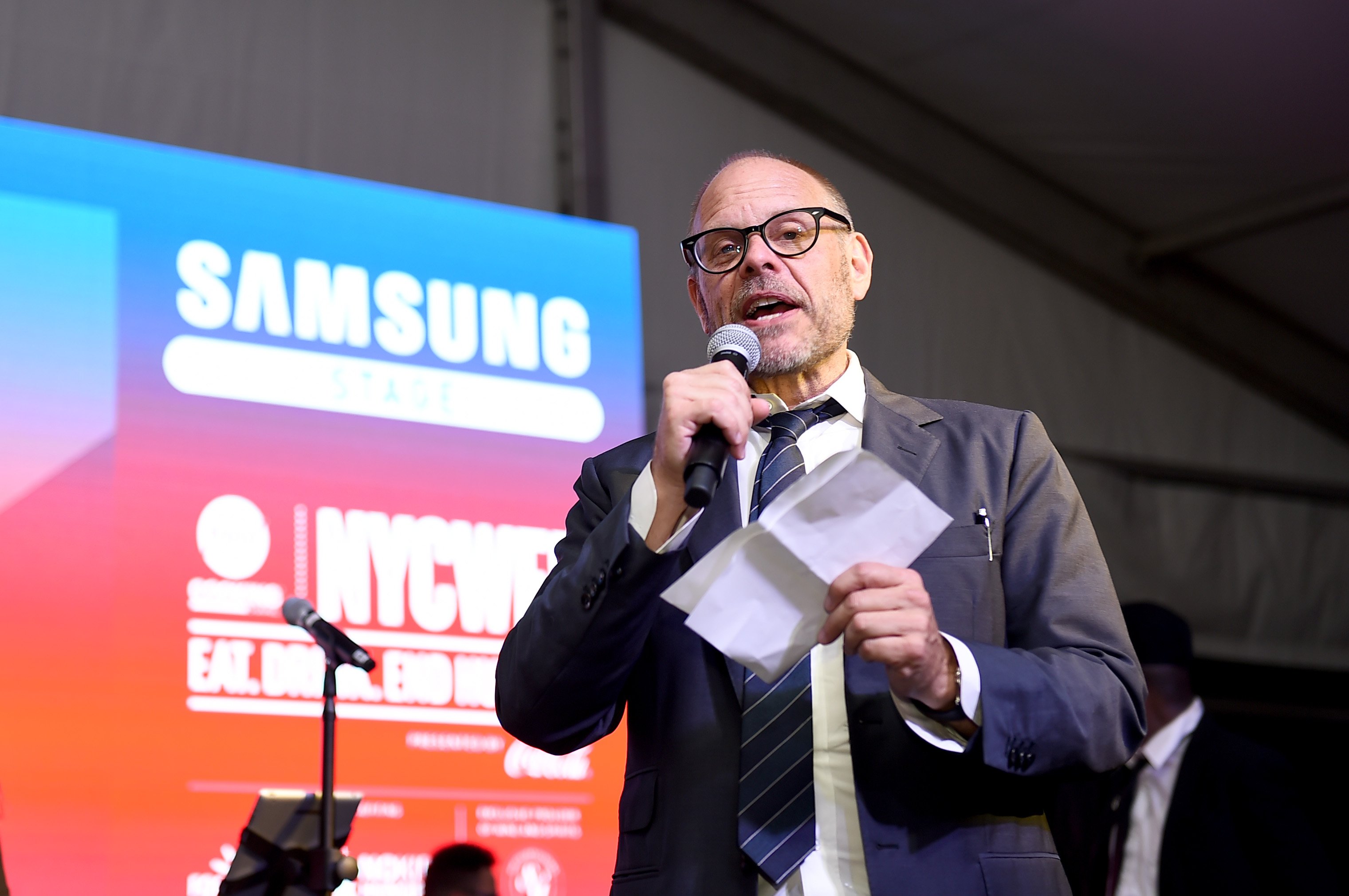 Culinary personality Alton Brown addresses an audience with a microphone in his hand.