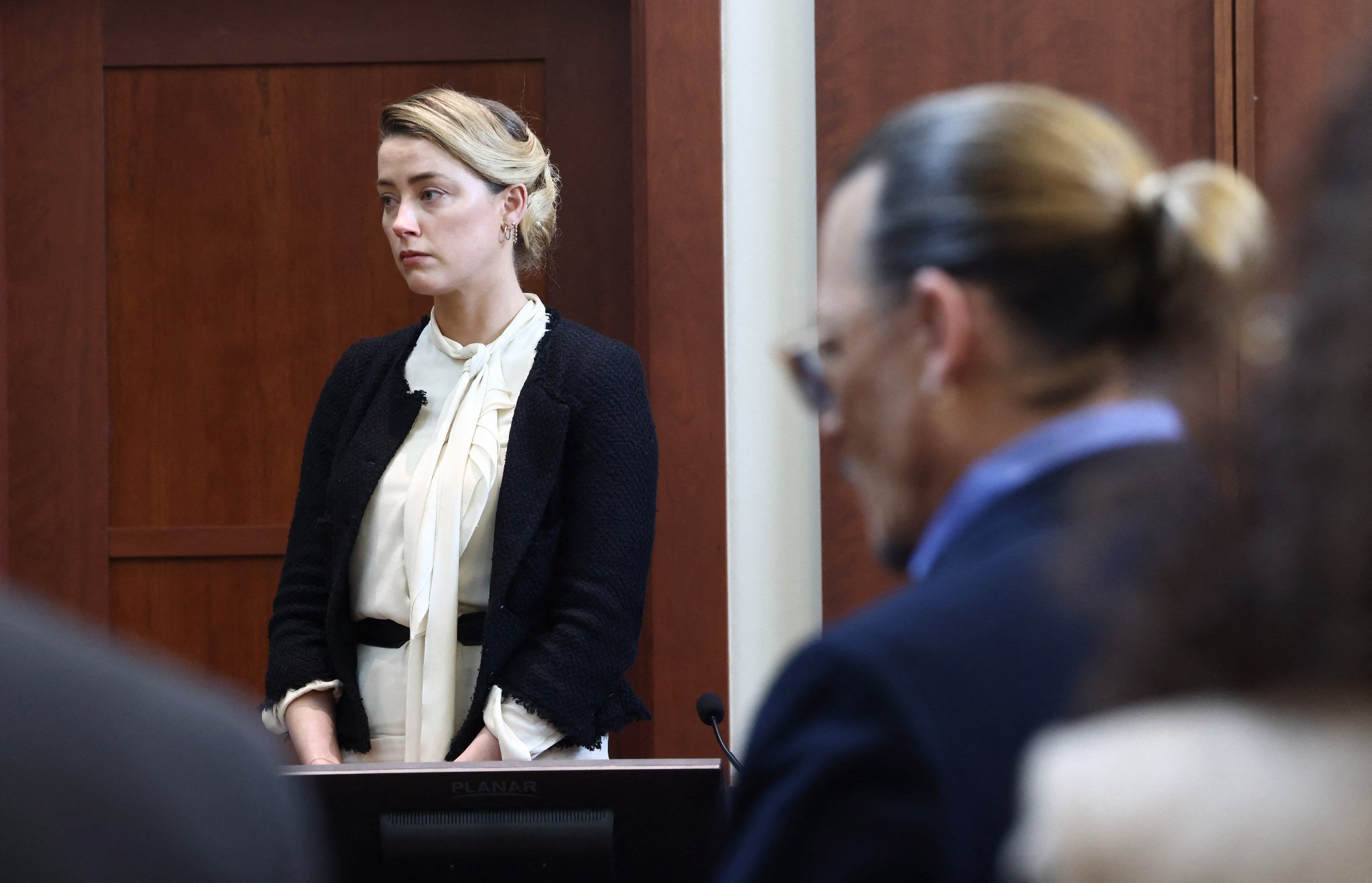 Amber Heard and Johnny Depp in court to testify for the jury to determine a winner in the defamation case. Heard is standing looking to the side and Depp is in the foreground with sunglasses on.