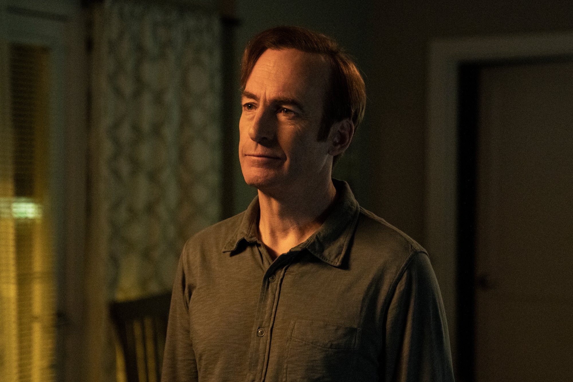 Bob Odenkirk as Saul Goodman in 'Better Call Saul' Season 6 Episode 7, which kills off Howard Hamlin. He's standing in a dimly lit room and wearing a brown shirt.