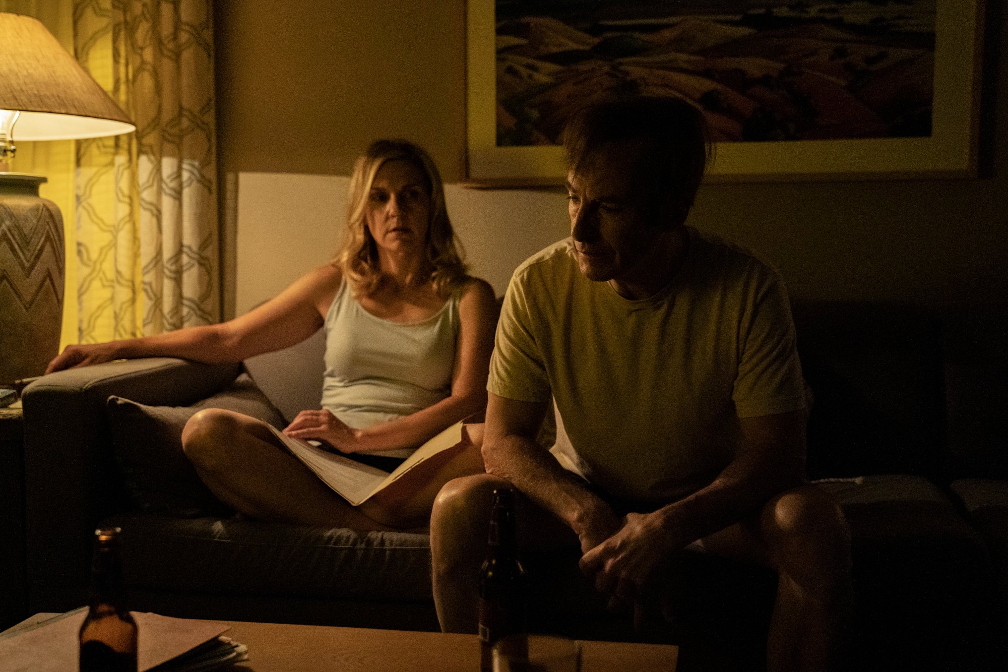 Rhea Seehorn and Bob Odenkirk as Kim Wexler and Jimmy McGill in 'Better Call Saul' Season 6 Episode 5, 'Black and Blue.' They're sitting on the couch in their dimly lit living room. Jimmy is leaning forward, and Kim is leaning back and looking at him.