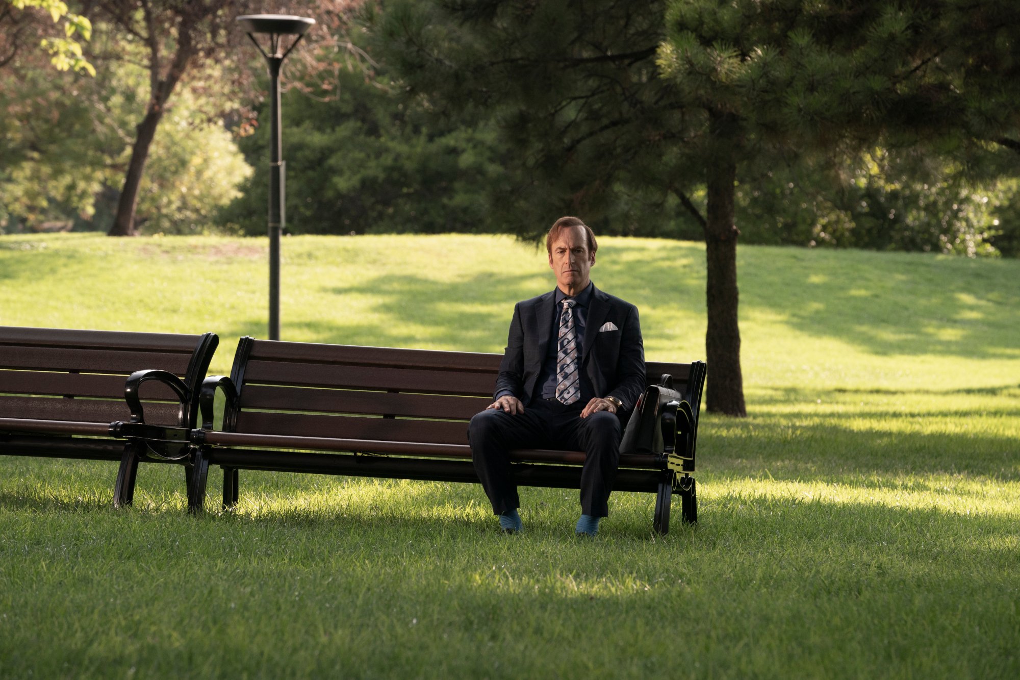 Bob Odenkirk as Saul Goodman/Jimmy McGill in 'Better Call Saul' Season 6 Episode 7, which is the final chapter's mid-season finale. He's sitting on a bench in a park, with grass and trees everywhere.