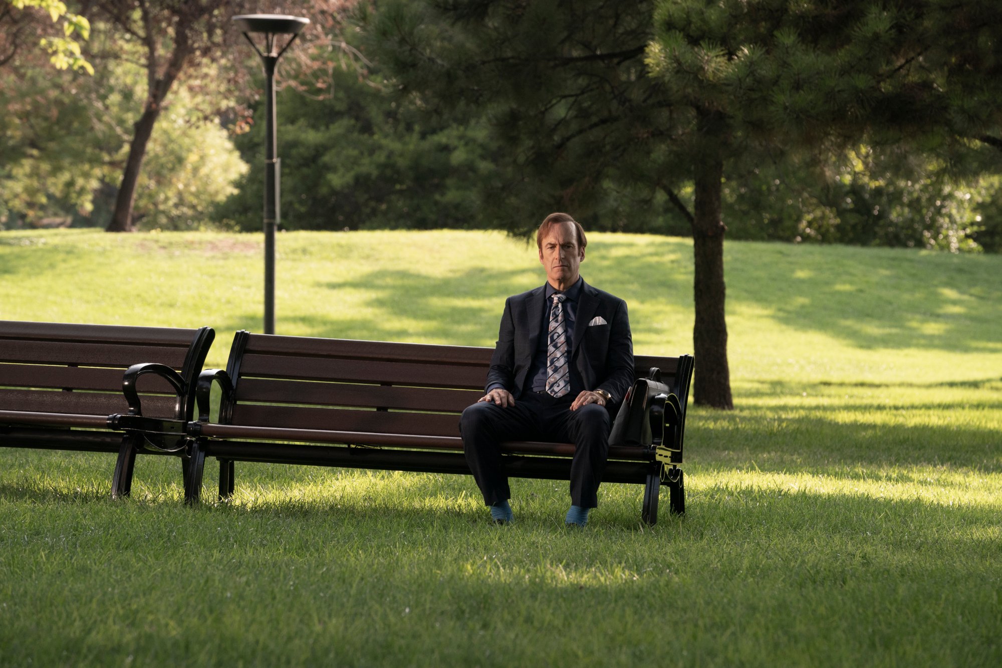 Bob Odenkirk as Saul Goodman/Jimmy McGill in 'Better Call Saul' Season 6 Episode 7, which is the final chapter's mid-season finale. He's sitting on a bench in a park, and there's grass and trees everywhere.