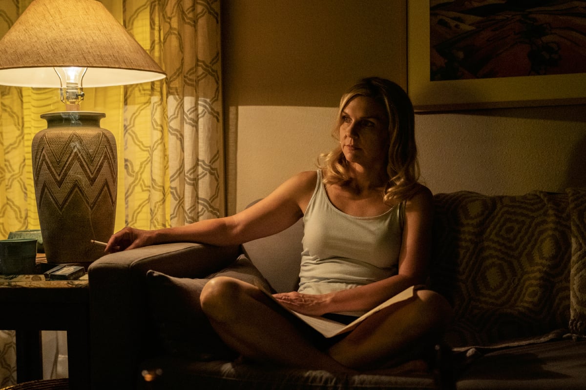 Rhea Seehorn as Kim Wexler in Better Call Saul Season 6. Kim sits on the couch with her legs crossed wearing a white tank top and holding a cigarette. 