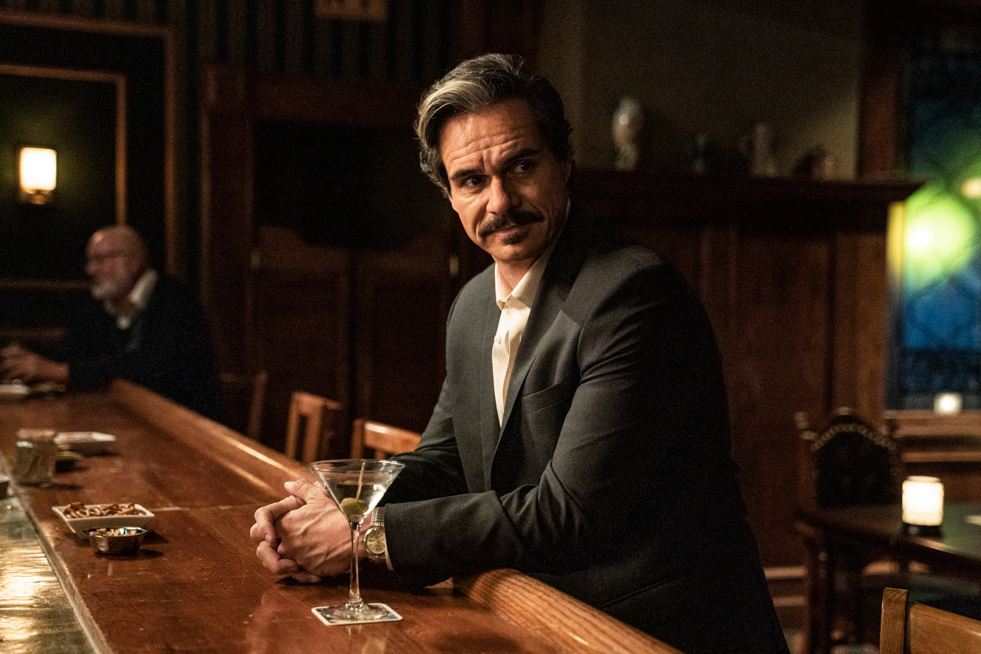 Tony Dalton as Lalo Salamanca in 'Better Call Saul' Season 6 Part 1, which leaves him wanting revenge against Gus in part 2. He's leaning against a bar, wearing a black suit, and smiling.