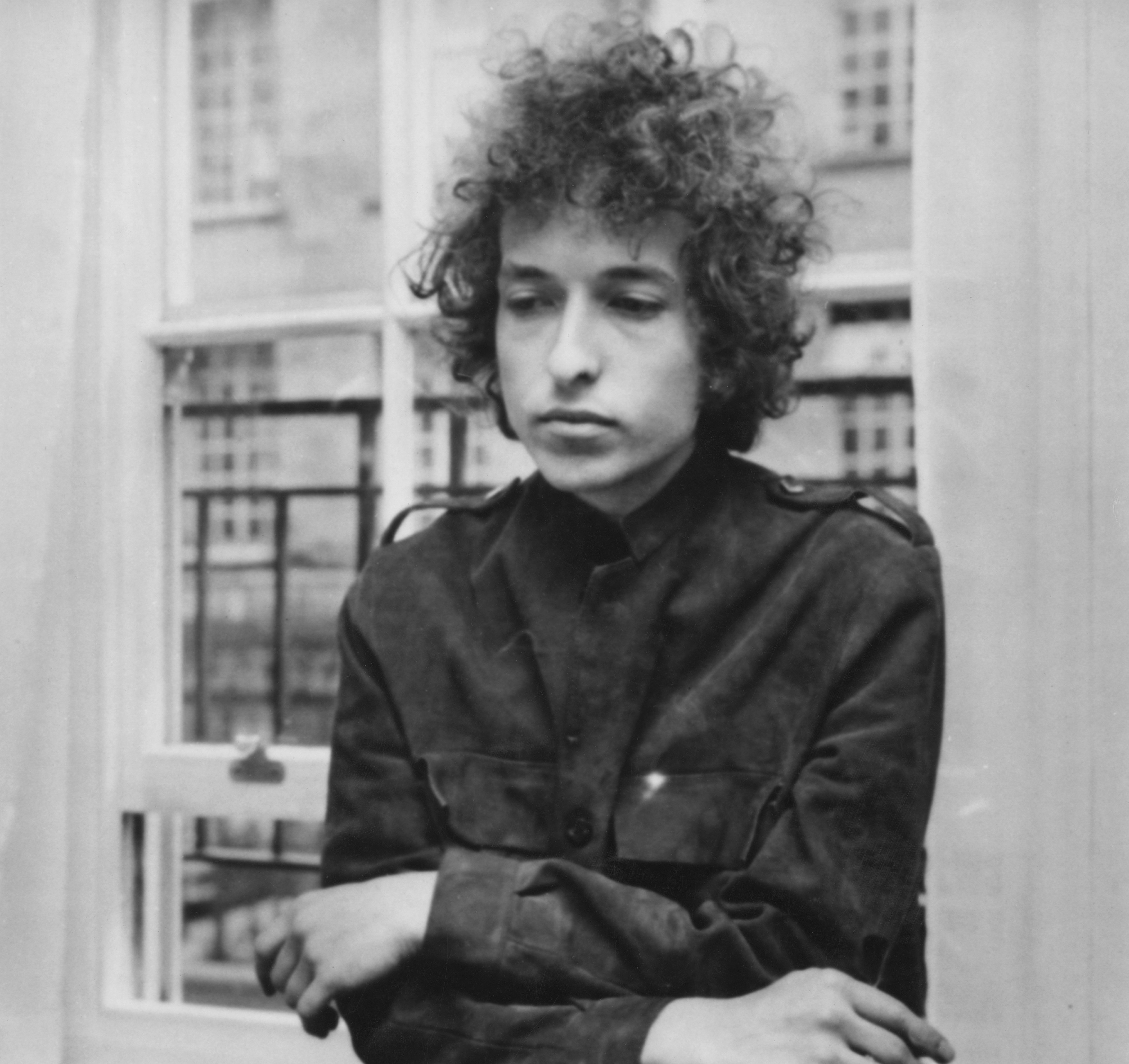 A black and white picture of Bob Dylan standing in front of a mirror with his arms folded.